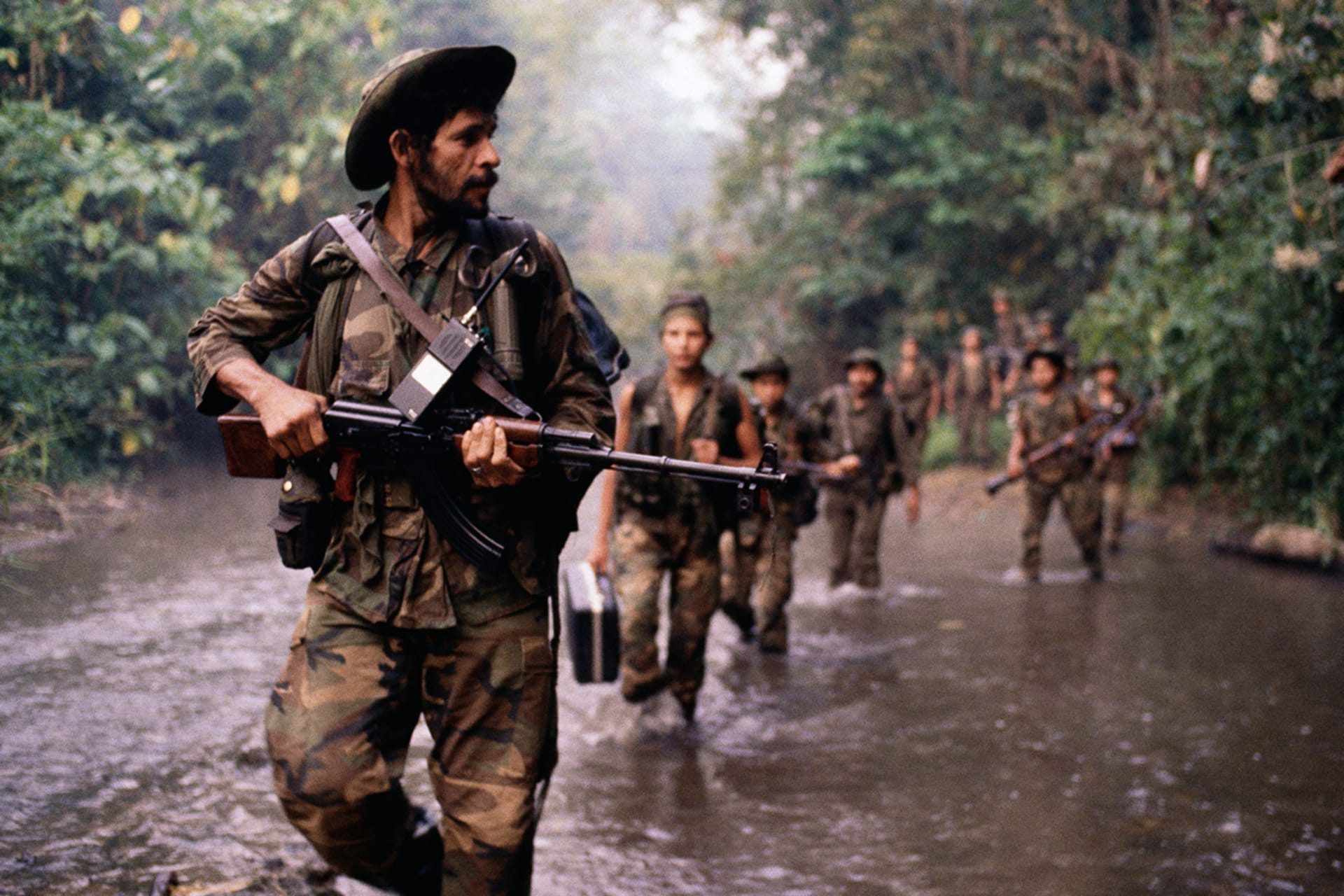 Men in military fatigues march with rifles through a waterway in a jungle.