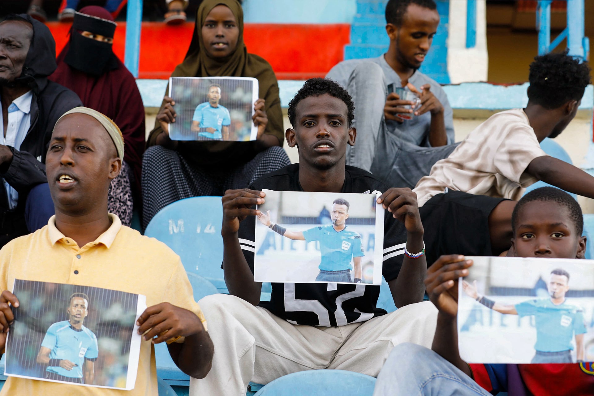 Somali soccer fans sit in a stadium, holding photos of referee Omar Abdulkadir Artan, who had been expected to officiate matches at the 2026 FIFA World Cup but was refused entry to the United States.