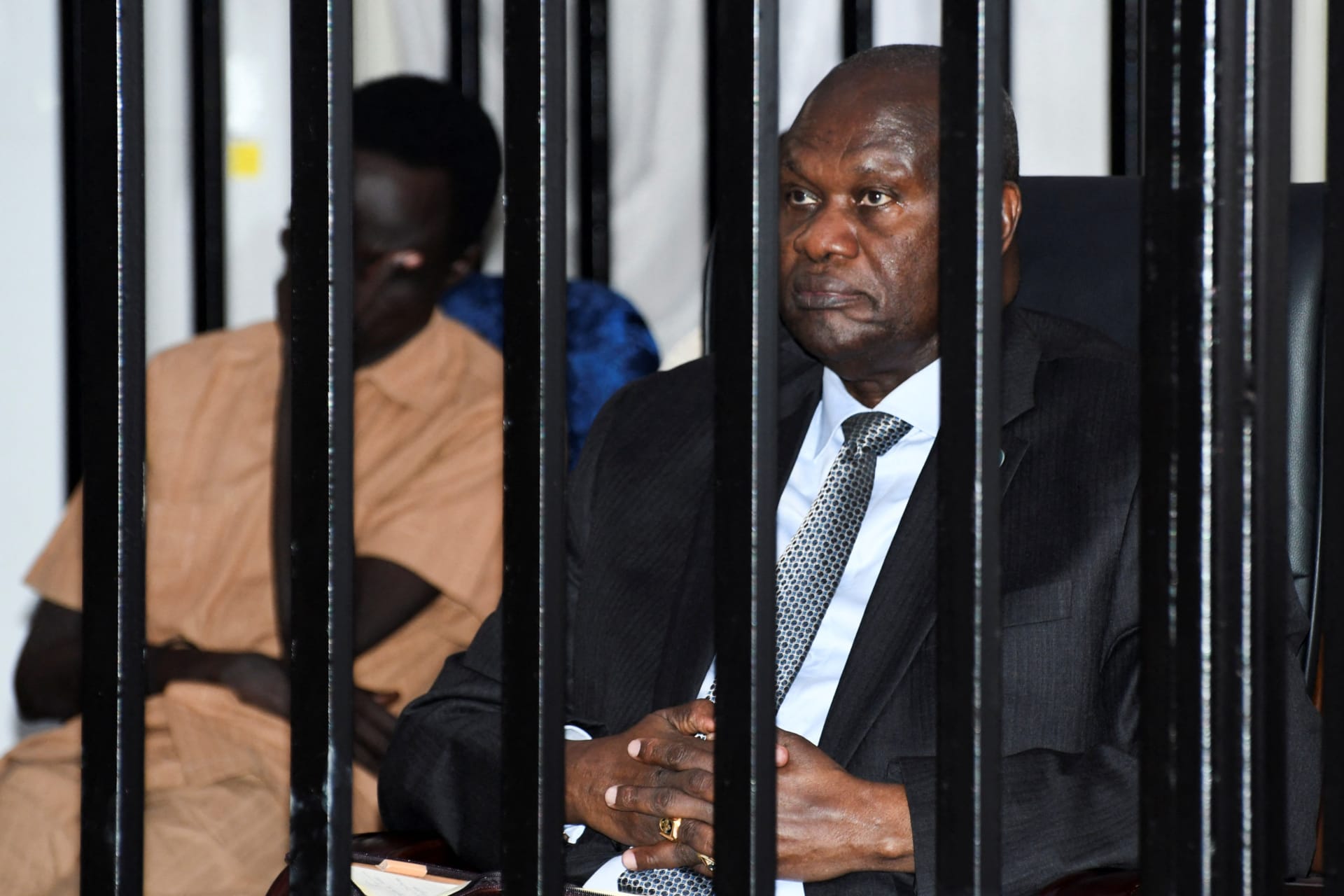 A man wearing a suit sits inside a black steel cage.