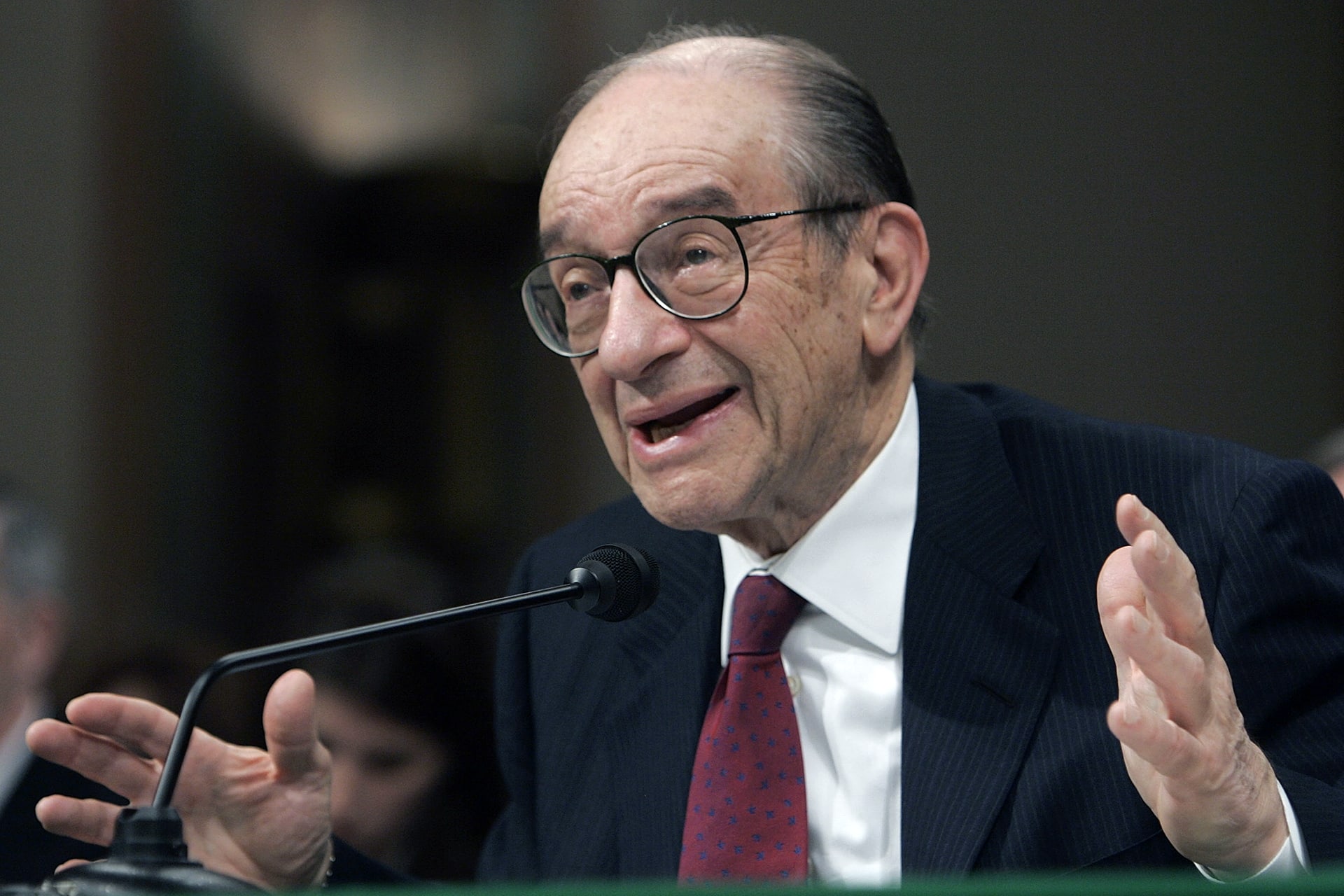 Alan Greenspan, dressed in a suit and red tie, gestures while speaking before a microphone.