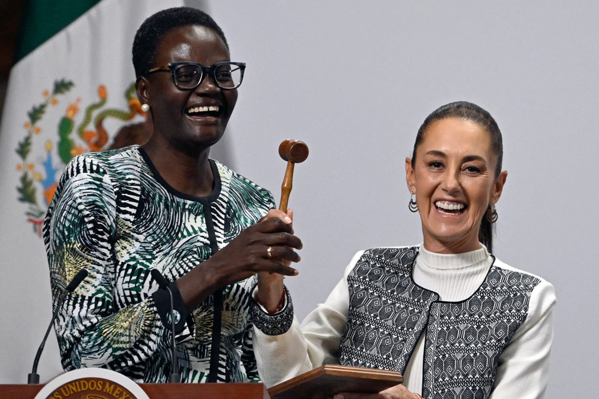 Tulia Ackson, left, stands and holds a gavel next to Claudia Sheinbaum, right, who holds the gavel block. The two smile and stand behind a wooden podium.