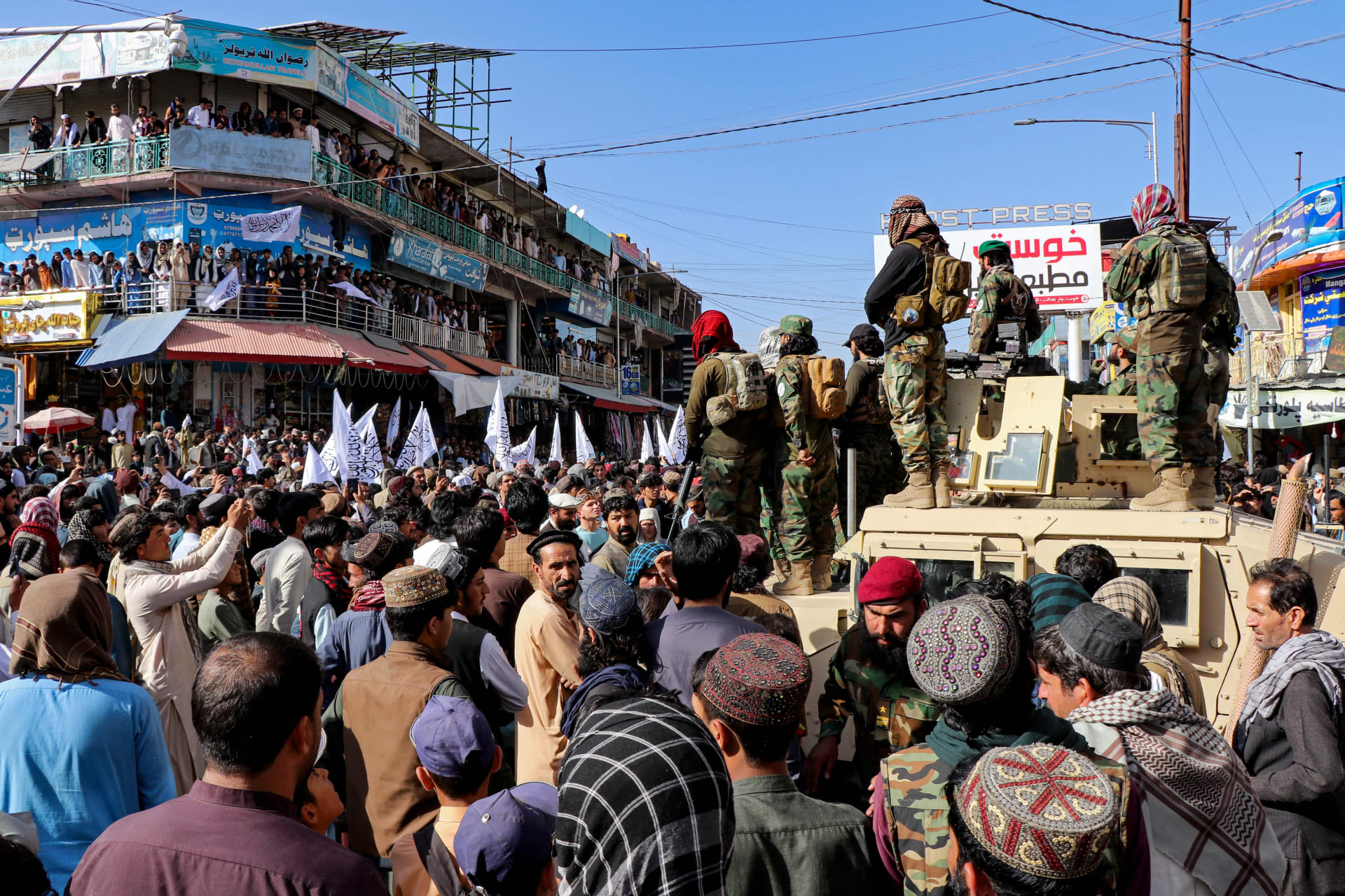 A crowd of many people stand in an urban area facing toward men on tanks.