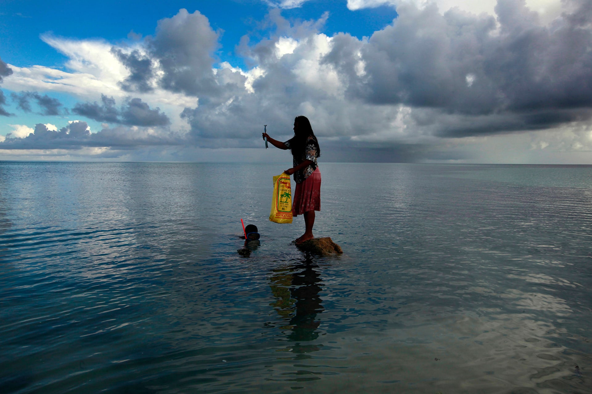 A woman stands on top of a rock holding a fish as her husband wears a snorkel and dives under the water to catch more fish.