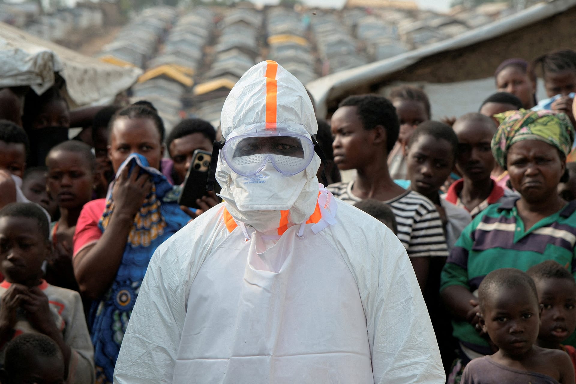 A health worker in a white hazmat suit stands in front of a group of people looking on in a displacement camp.
