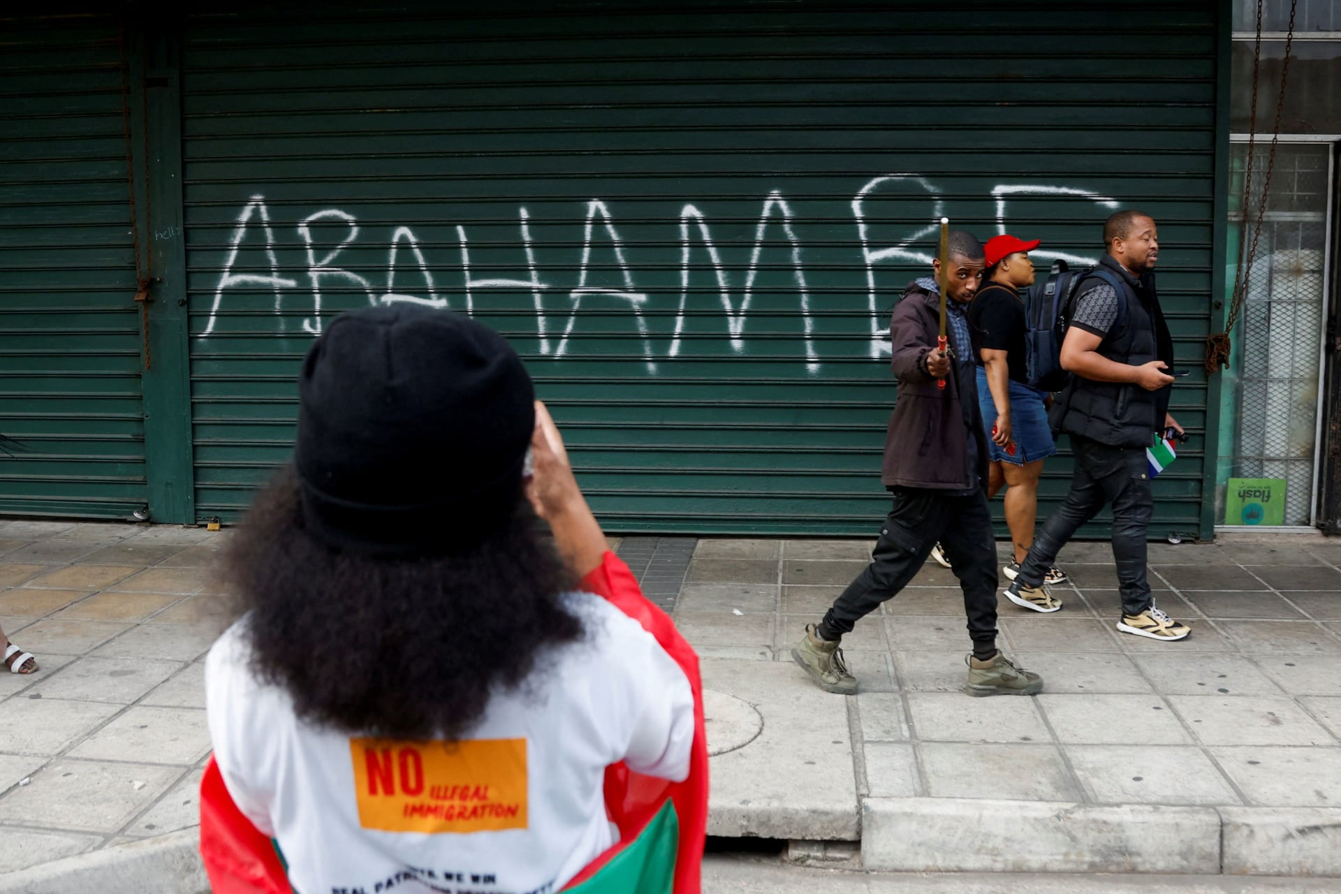 <p>People walk past a slogan reading, “They must go,” written on the shutter of a shop as anti-immigrant protesters march on the day of an unofficial deadline set by anti-immigrant groups for all undocumented migrants to leave, in Durban, South Africa, on June 30, 2026. </p>
