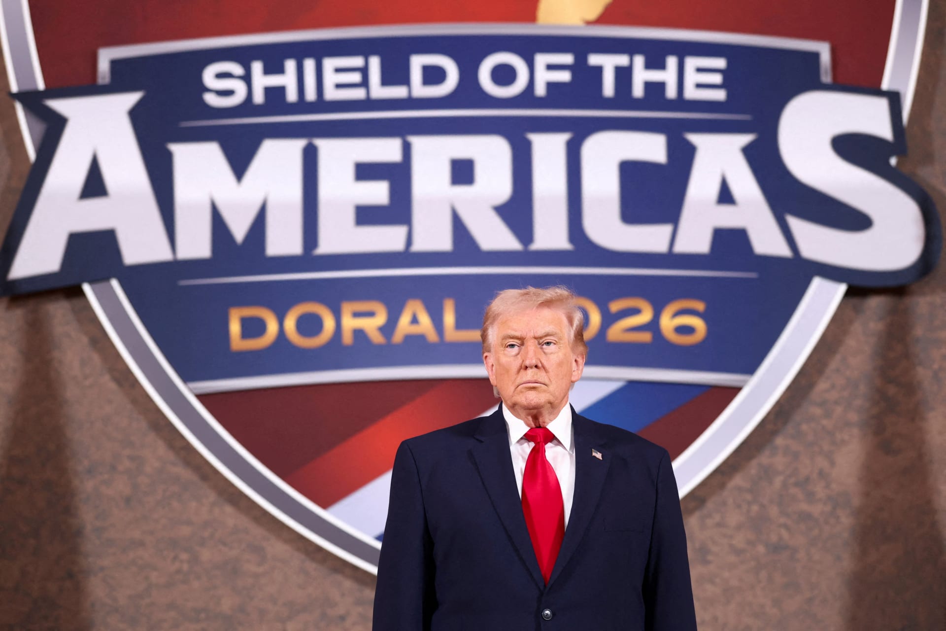 Donald Trump stands in front of a Shield of the Americas logo