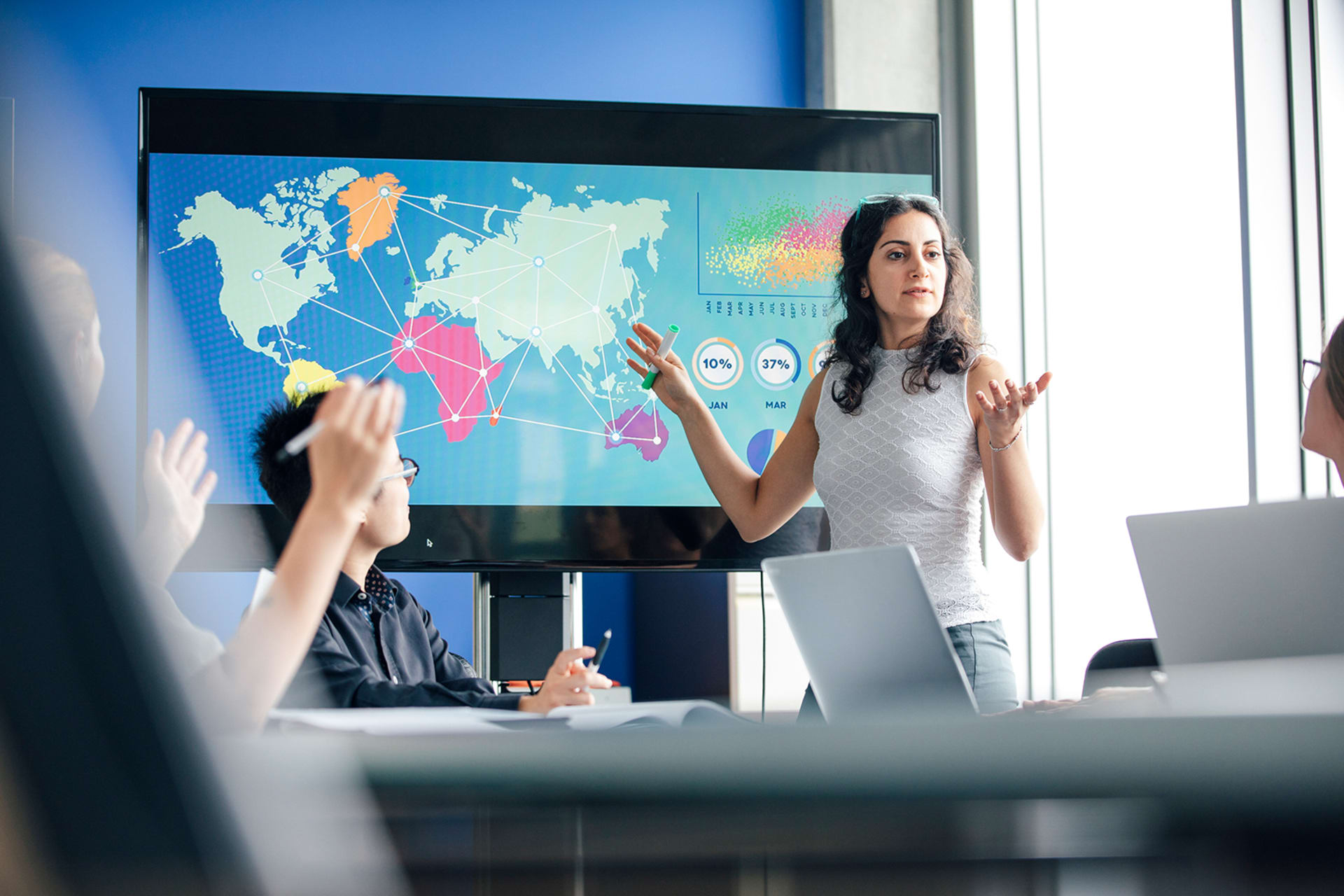 A woman stands in front of a screen that shows a world map. Other are seated at a table listening to her.