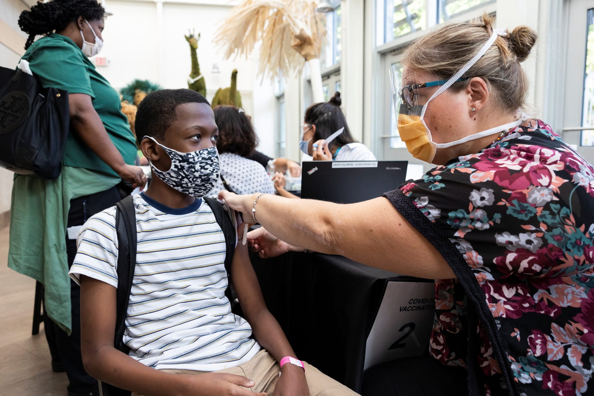 A boy sits in a chair while getting a vaccine from a health worker, both of them wear face masks.