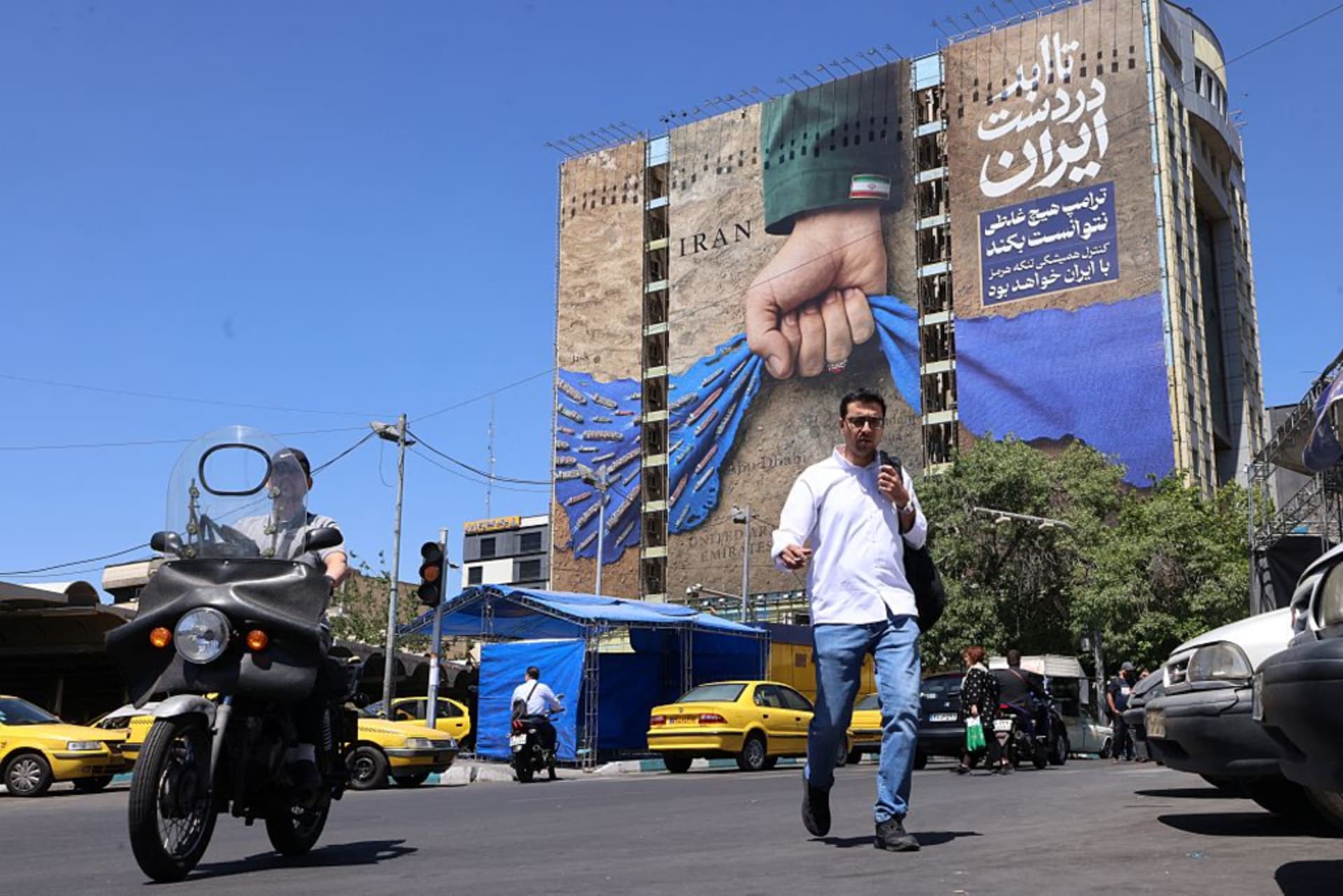 People cross a street past a billboard on the facade of a building depicting the Strait of Hormuz with a caption in Persian reading "Forever in Iran's Hand", at Vanak Square in Tehran on May 25, 2026. I