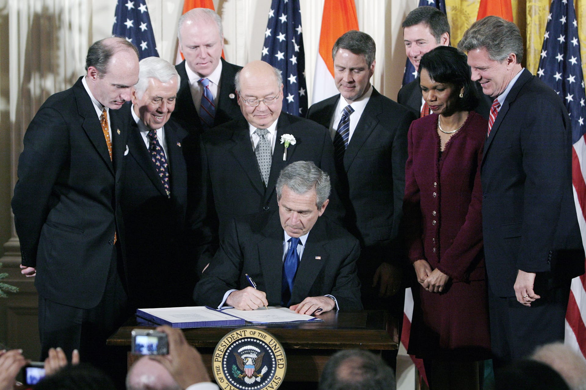 George W Bush signs a document while surrounded by key advisors and members of Congress.
