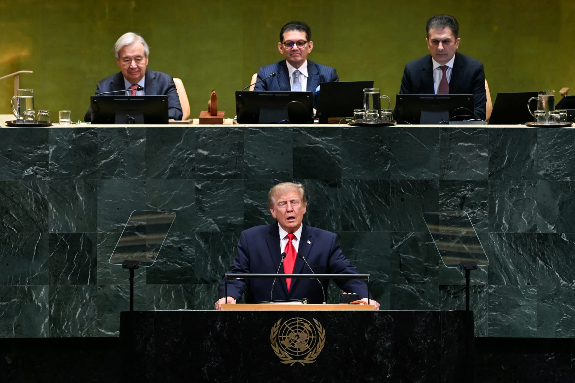 Trump stands at the green marble General Assembly rostrum, three UN officials seated at the dais above and behind him.