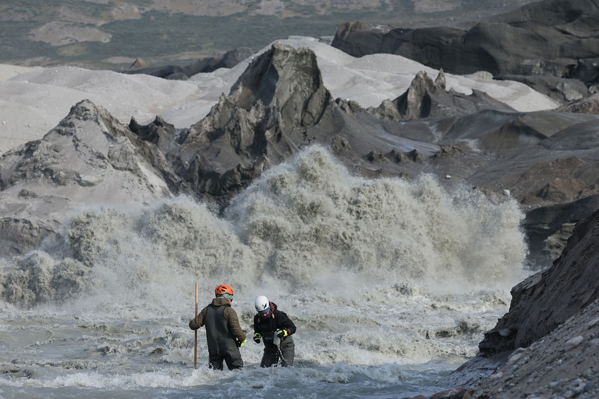 Scientists in gear collect glacial meltwater and sediment samples. There is rapidly moving water around them.