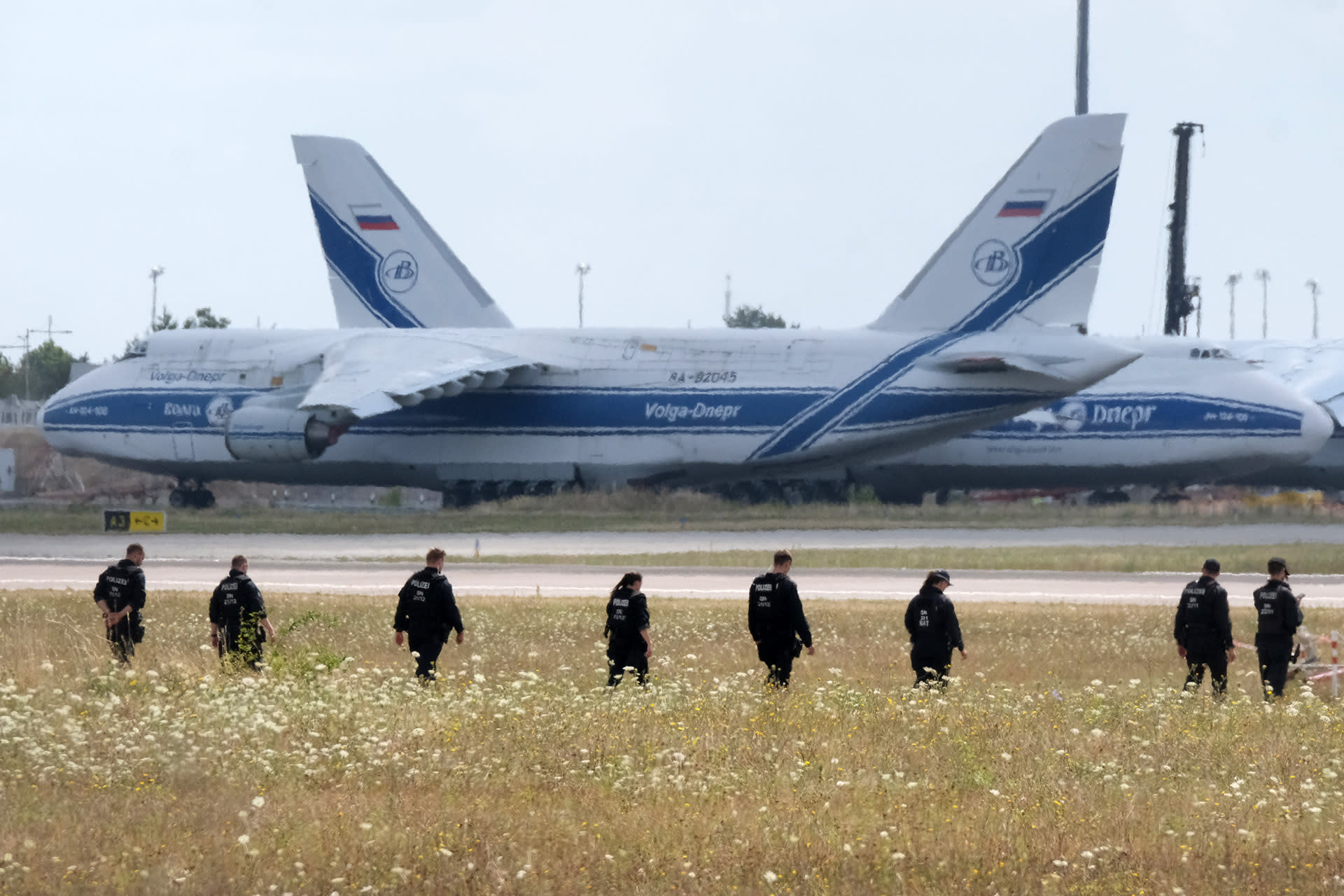 A line of police officers walks through a field of tall grass toward a runway at Leipzig Airport.
