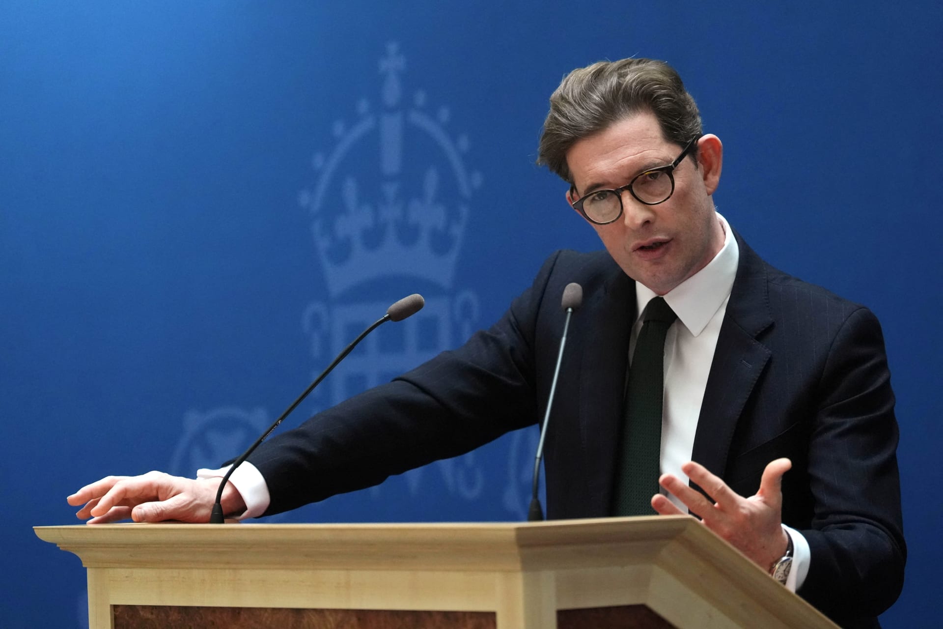 Ken McCallum speaks at a lectern with two microphones against a blue backdrop marked with a royal crest.