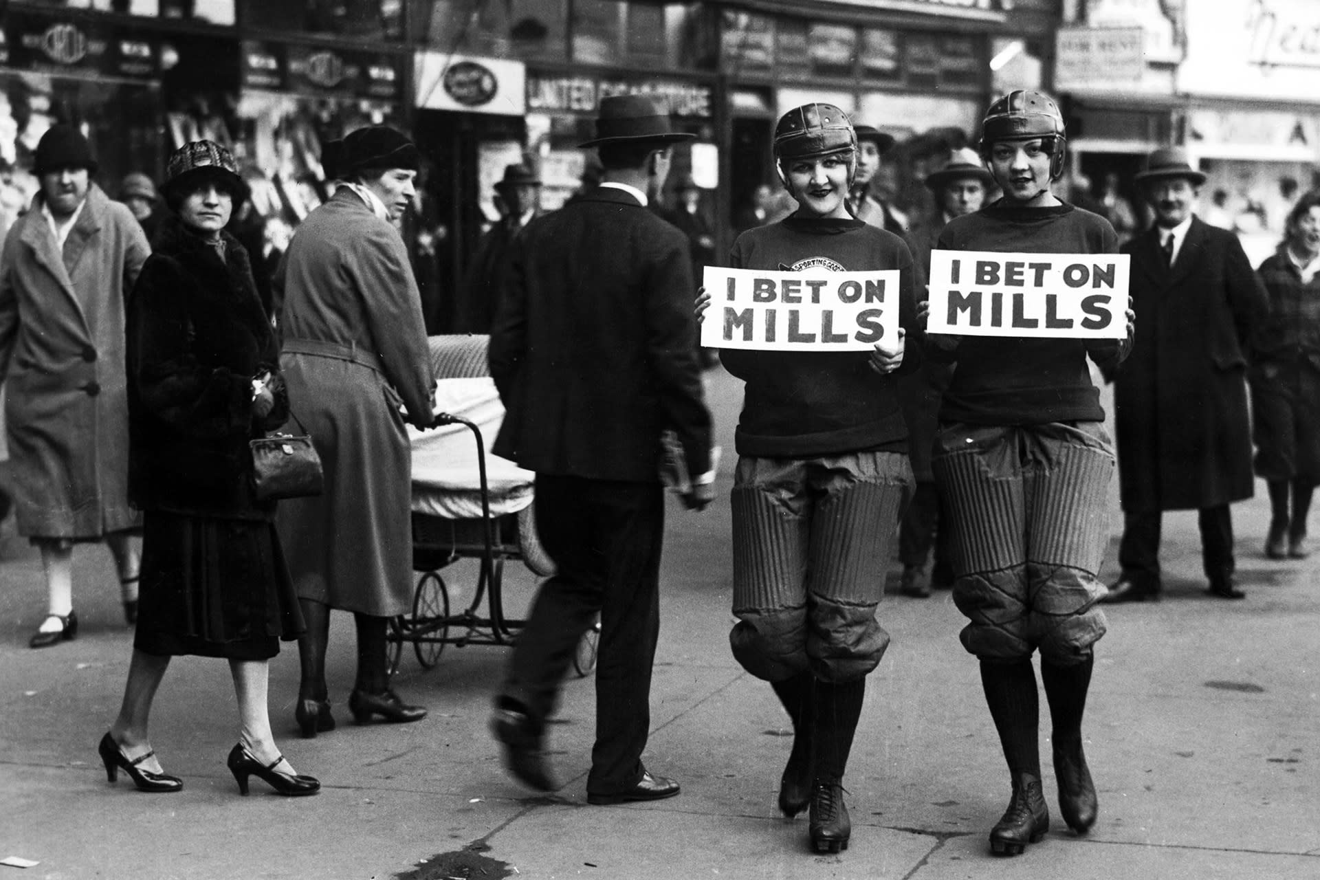 Two girls in football gear holdin signs that read "I Bet on Mills" walk on a crowded New York City street.