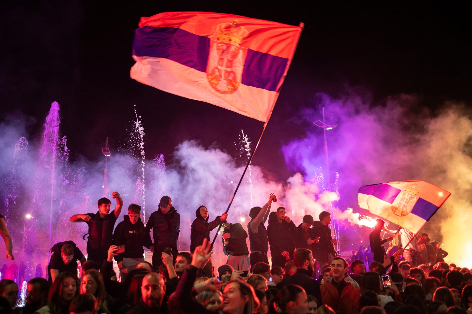 A crowd of people celebrates at night around an illuminated fountain in Belgrade’s Slavija Square, one of them waving a large Serbian flag amid colored smoke.