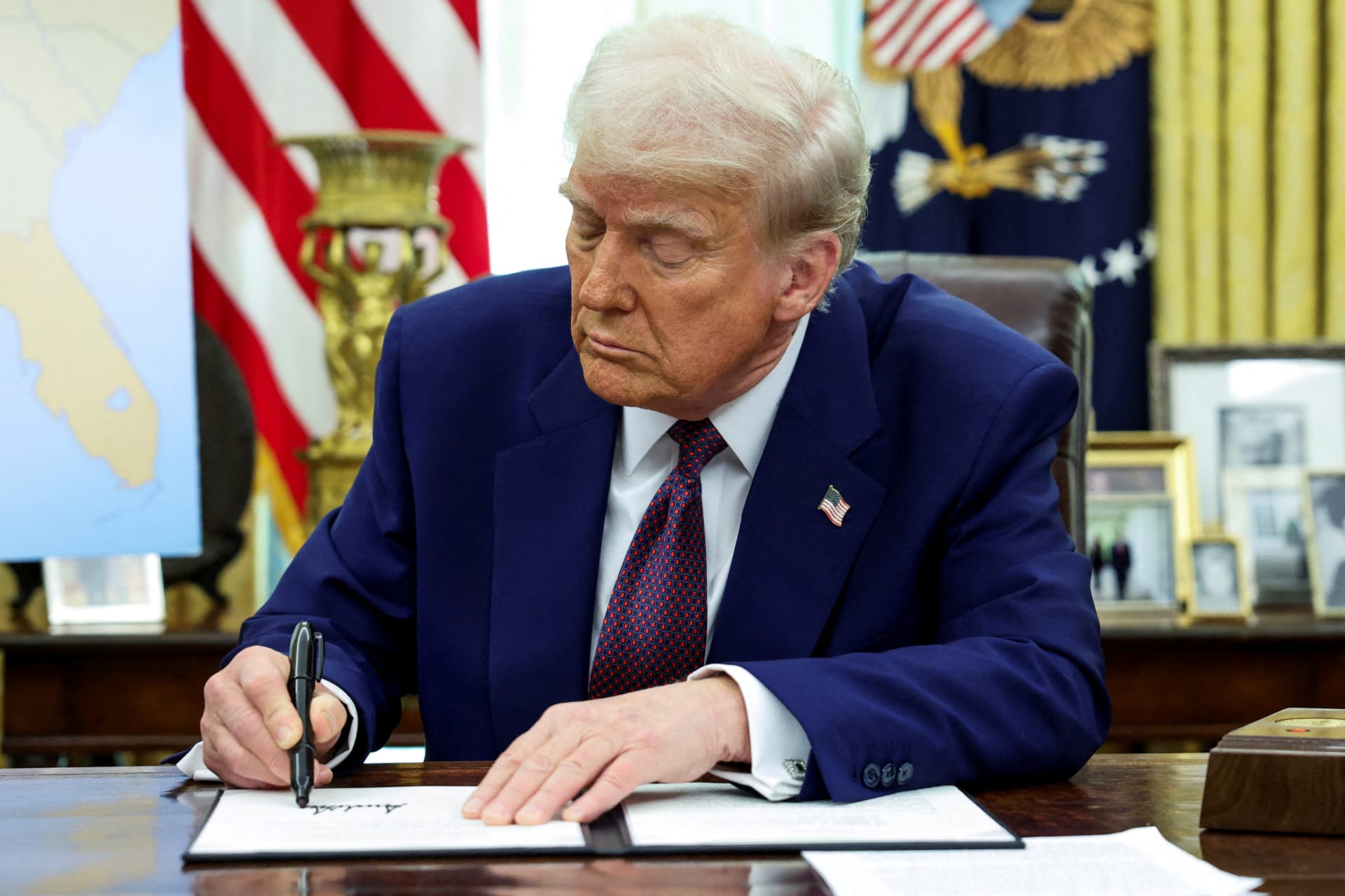 President Donald Trump signs an executive order at the Resolute Desk in the Oval Office, with an American flag and a map behind him.