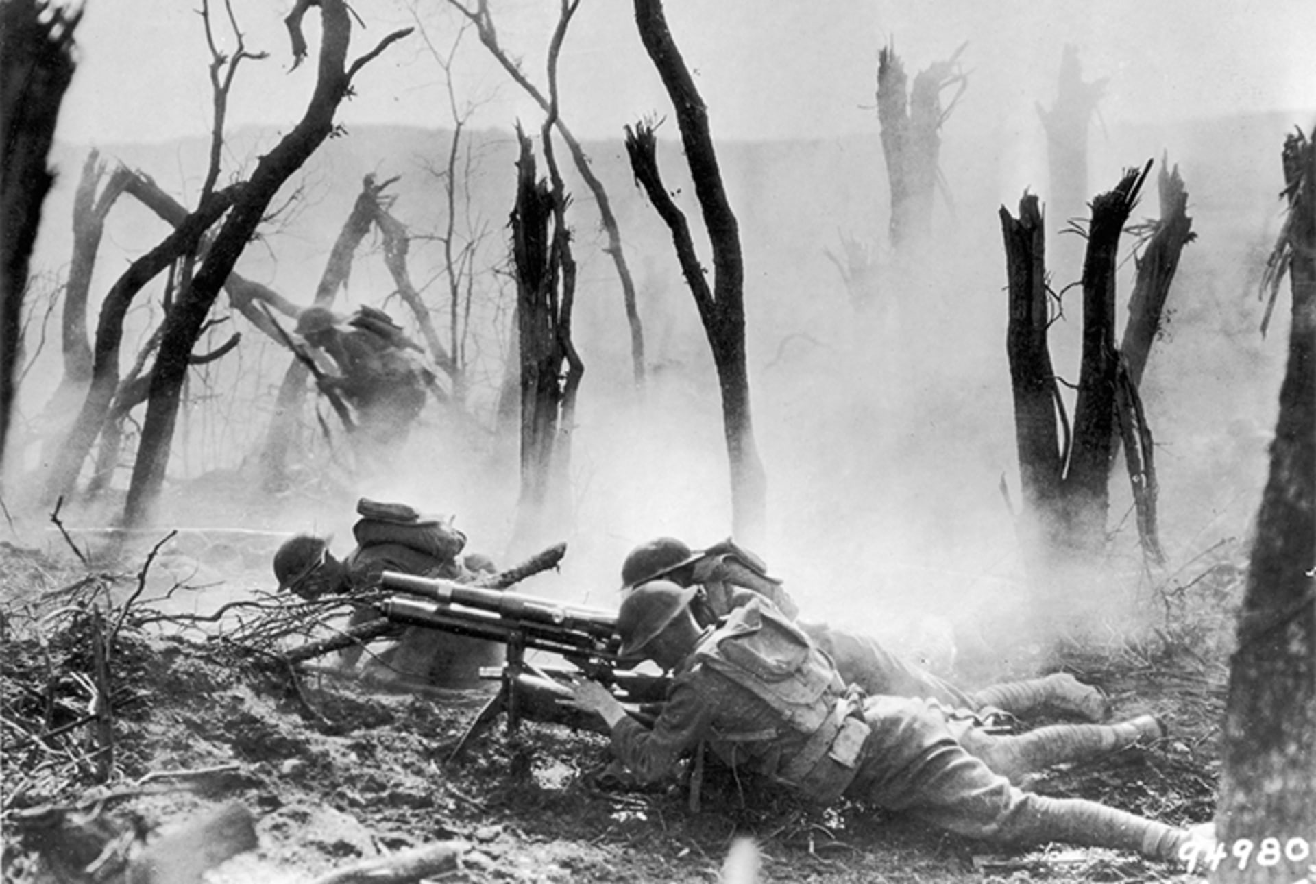 A black and white still from a documentary showing U.S. soldiers, surrounded by destroyed trees, laying down and firing a machine gun at German forces. 