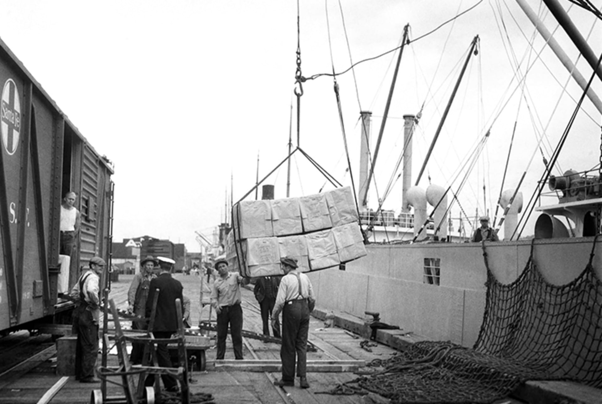 A black and white photo of longshoremen steadying a load of crates being lowered from a ship to the dock. A freight car stands to the left waiting to be loaded.