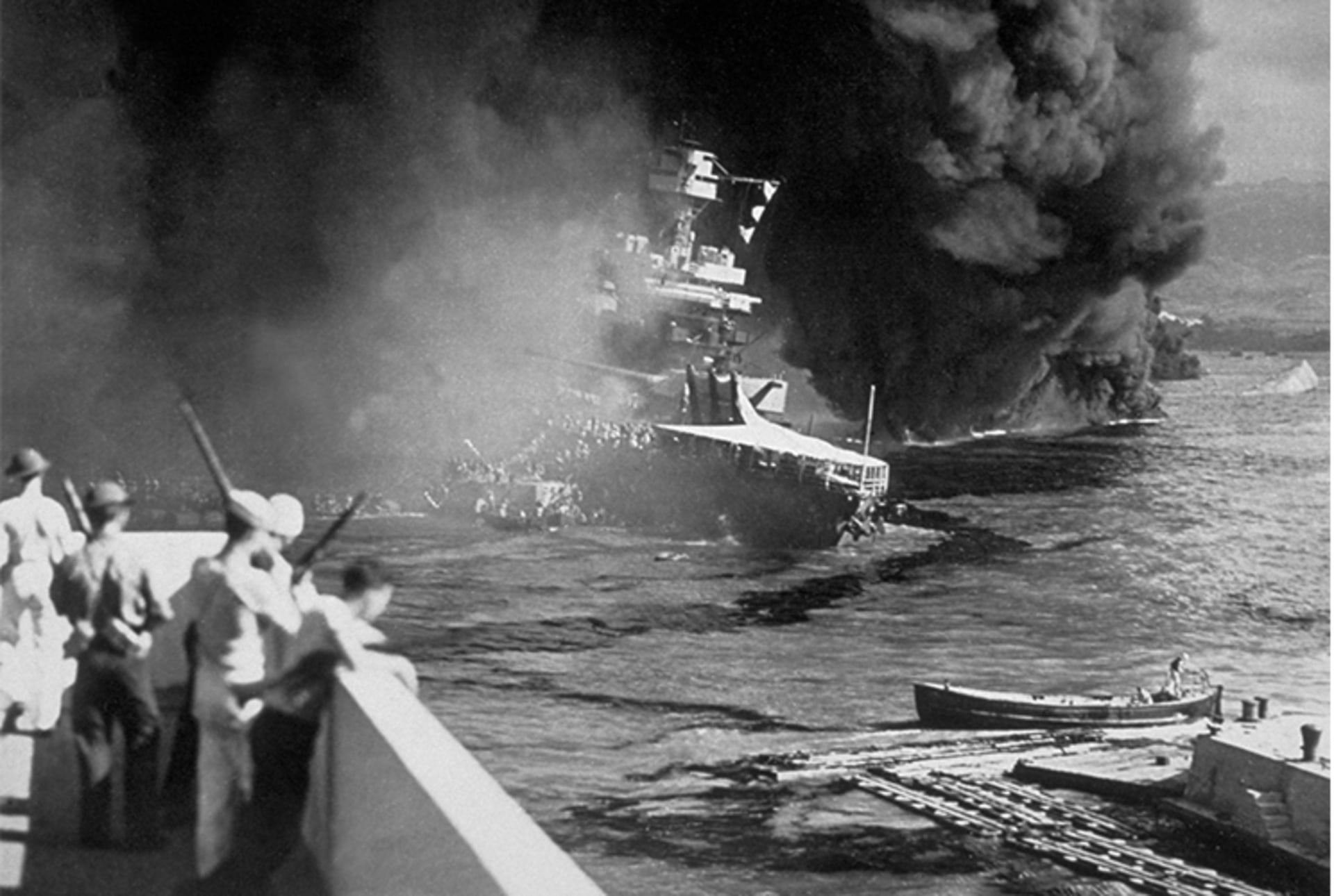 A black and white image of a U.S. ship on fire after the Pearl Harbor attack. Soldiers look on at the burning sight. 
