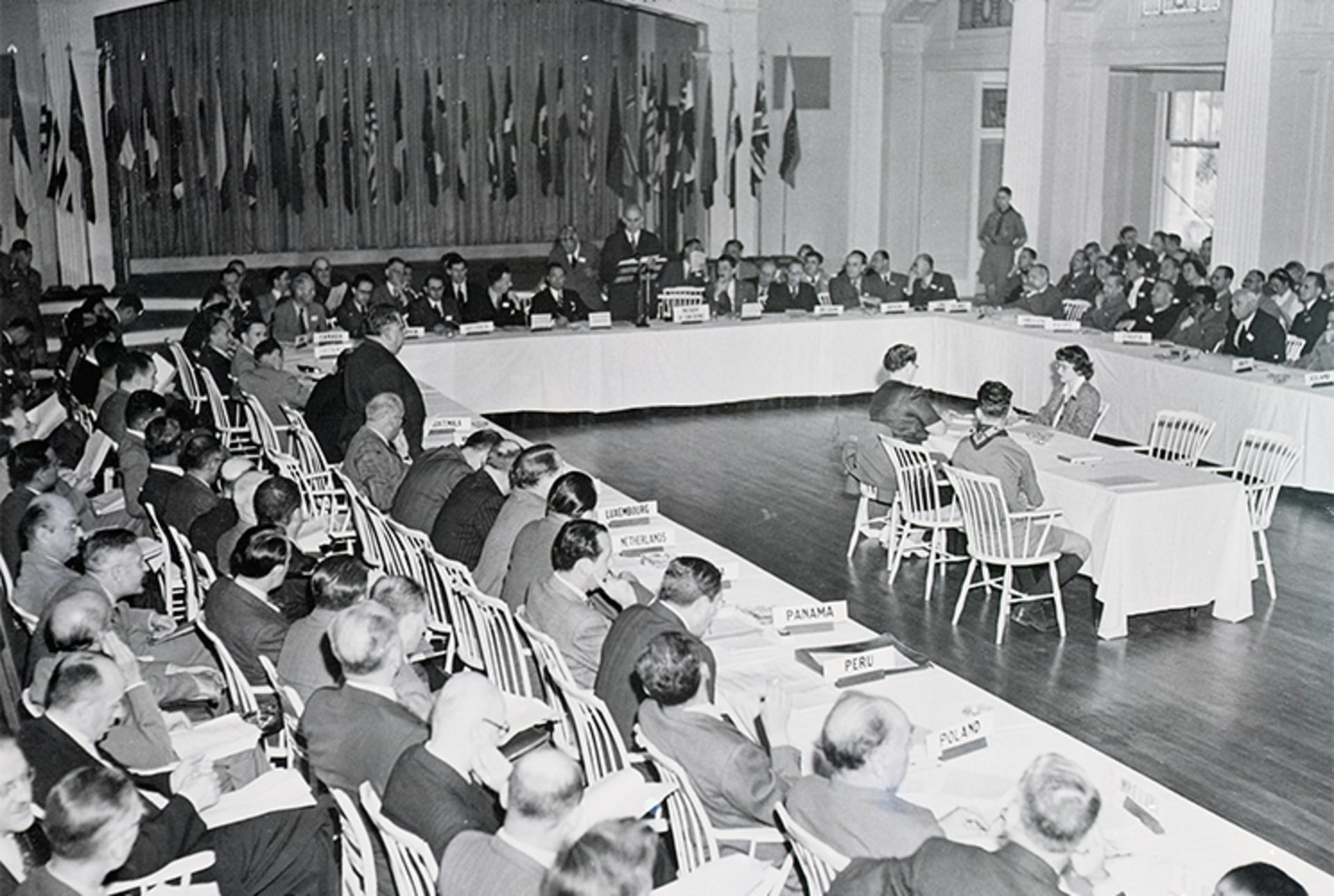 A black and white photo of members gathered at the Bretton Woods Conference. The background shows various United Nations country flags.