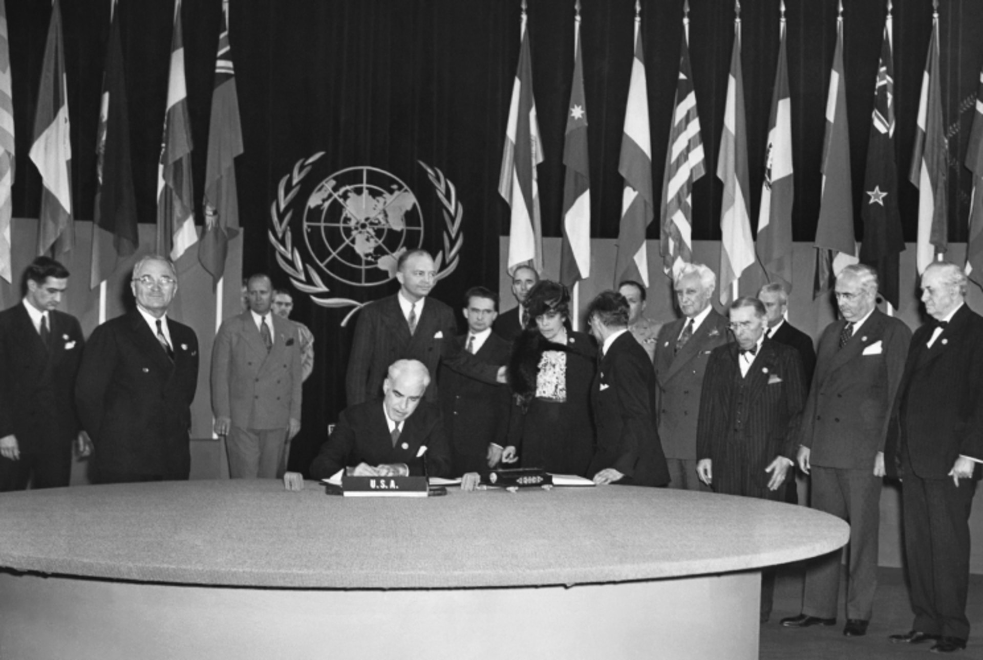 A black and white photo of the signing of the UN Charter. The UN logo and member country flags are in the back.