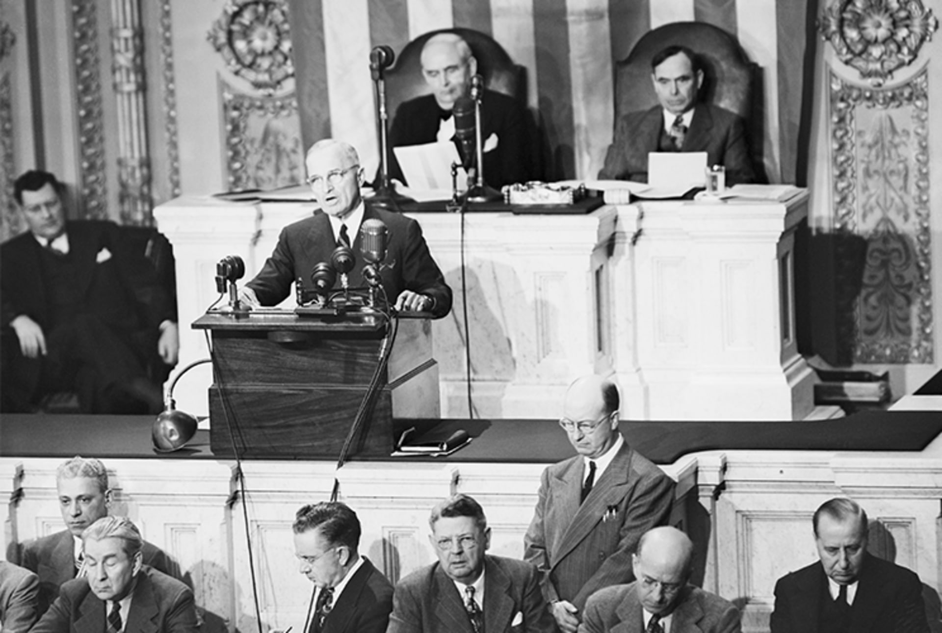 A black and white photo of President Truman addressing Congress from a podium with many mics. 