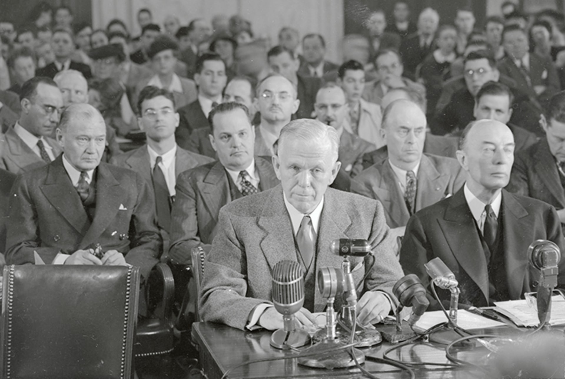 A black and white photo of Secretary of State George Marshall seated at a desk with many mics as he answers questions from the Senate.