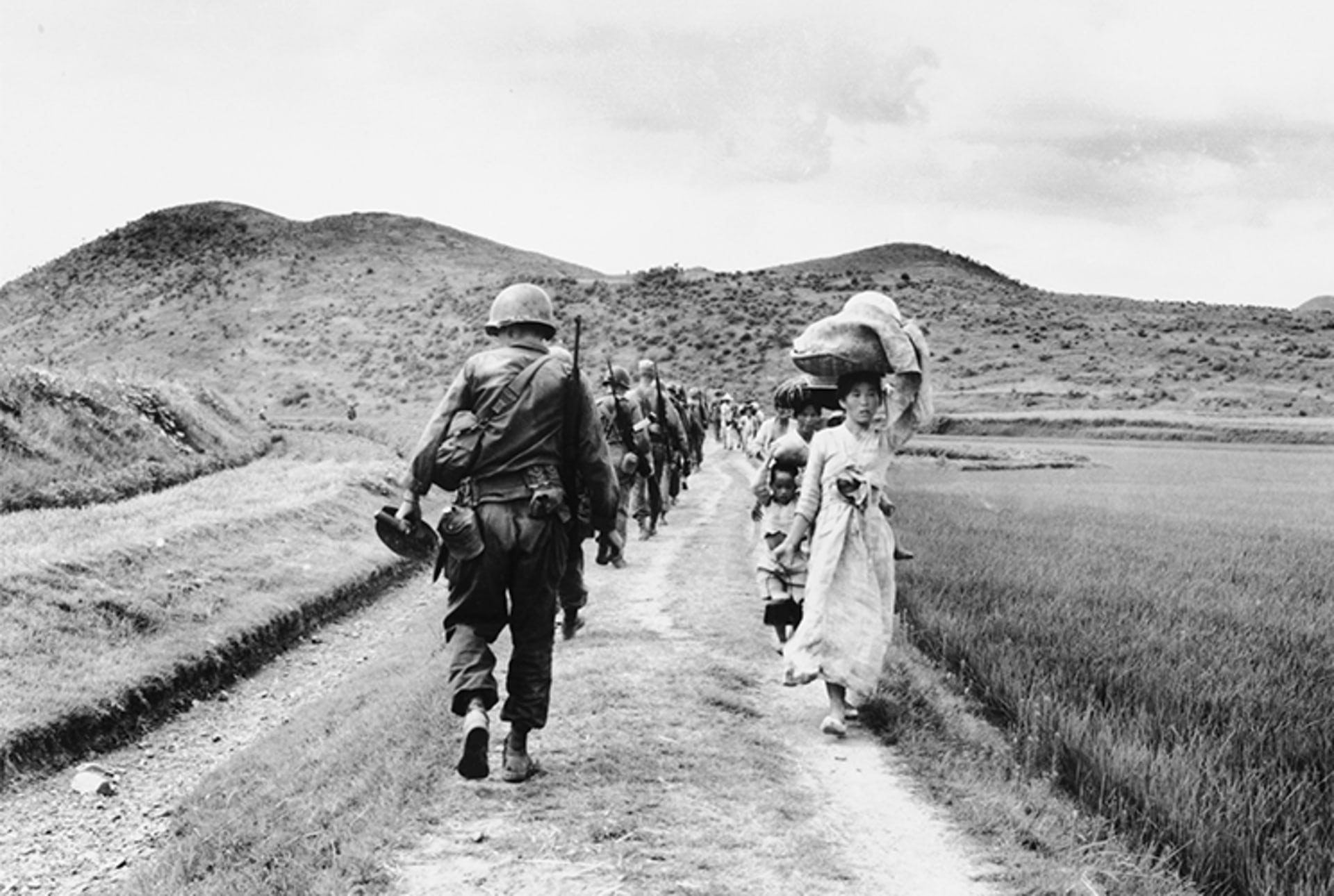 A black and white photo of a line of U.S. soldiers walking away from the camera on the left side and a line of Korean refugees walking towards the camera, carrying their belongings, on the right side.