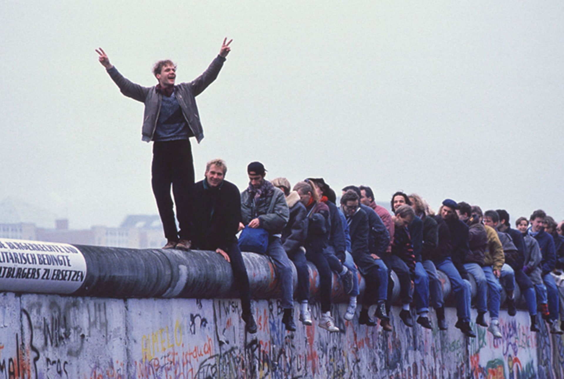 A photo of people atop the Berlin Wall celebrating the collapse of communism. The man at the front stands, while the rest of the group is seated, and raises his arms holding up two peace signs with his fingers. 