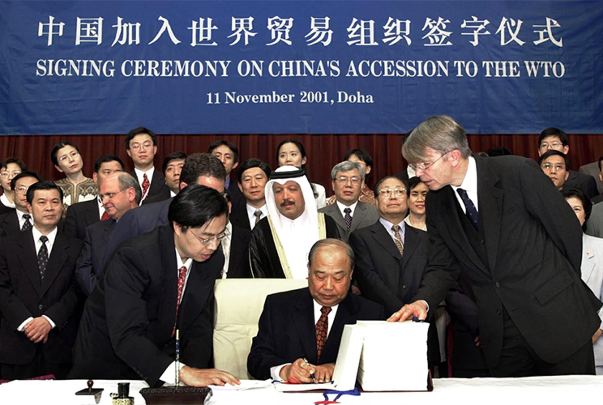A photo of Chinese Foreign Trade and Economic Cooperation Minister Shi Guangsheng signing China's WTO accession documents as people look on. There is a banner that reads "Signing Ceremony on China's Accession to the WTO" in the background in both Chinese and English. 