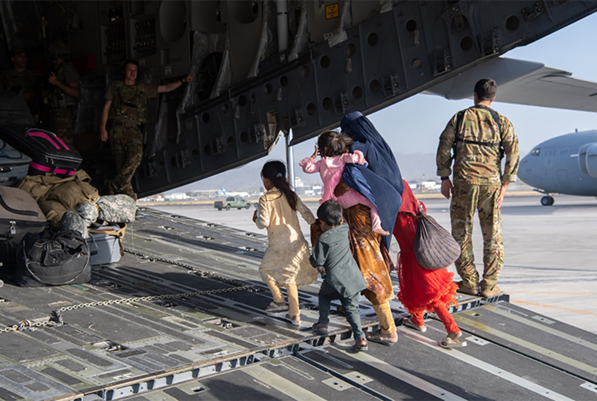 A photo of U.S. soldiers helping load passengers onto a U.S. plane at an airport as people evacuate. A woman with four kids walks on to the plan which has been loaded with many belongings. 