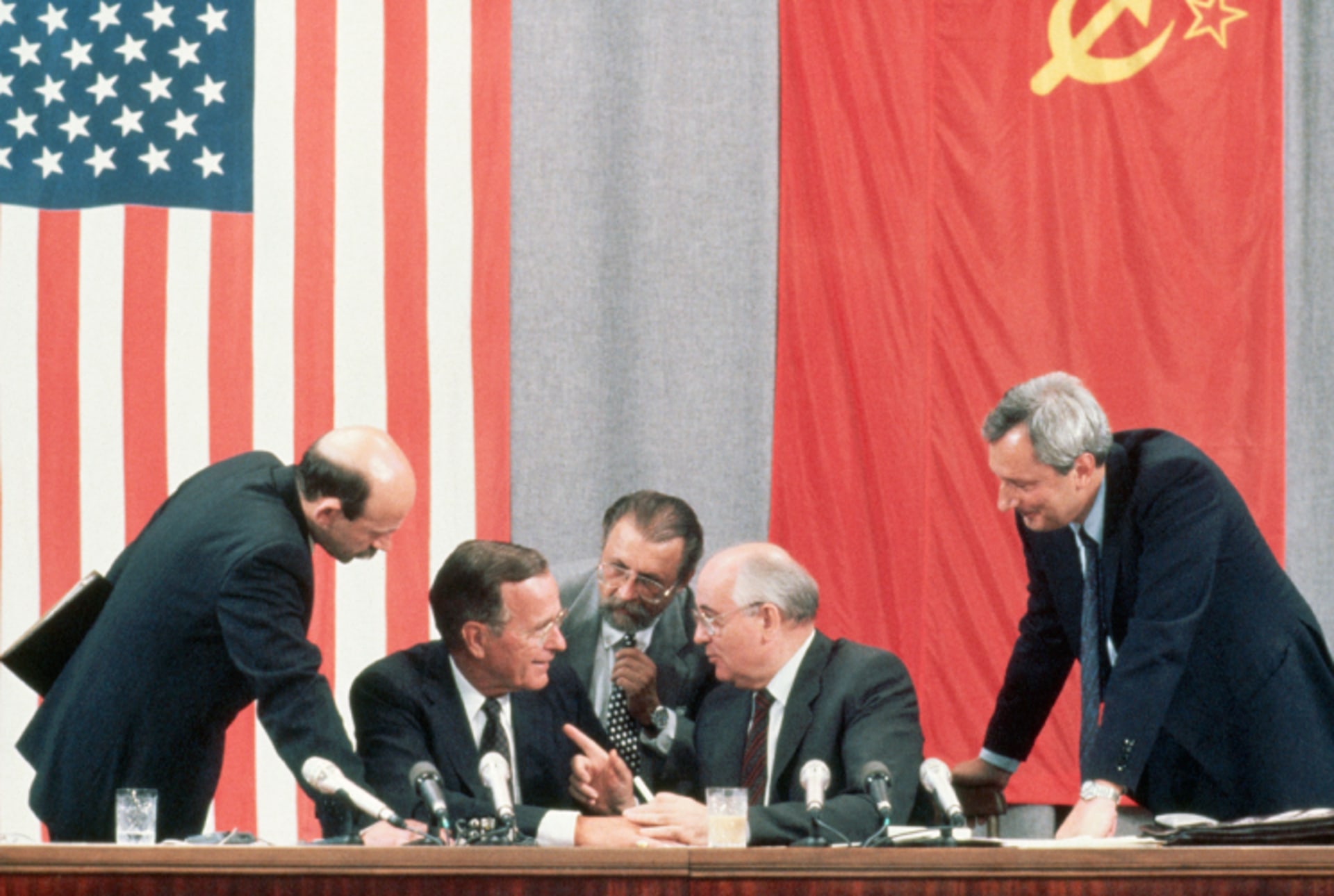 A photo of President George Bush and Soviet leader Mikhail Gorbachev during the 1991 Moscow Summit. The two converse as other men listen in. Behind them are the American and USSR flags hung on the wall.