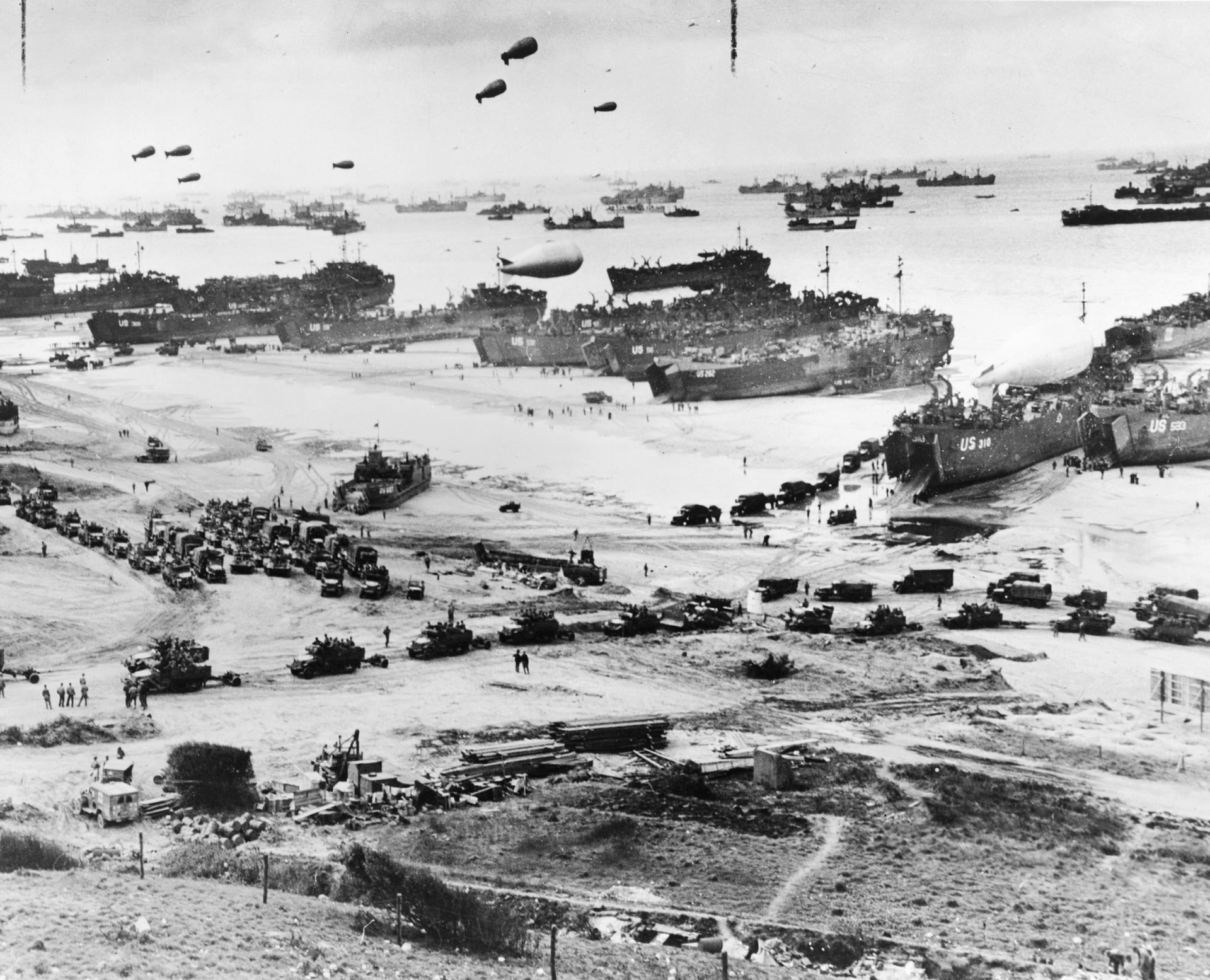 Bird's-eye view of landing craft, barrage balloons, and allied troops landing in Normandy, France on D-Day.