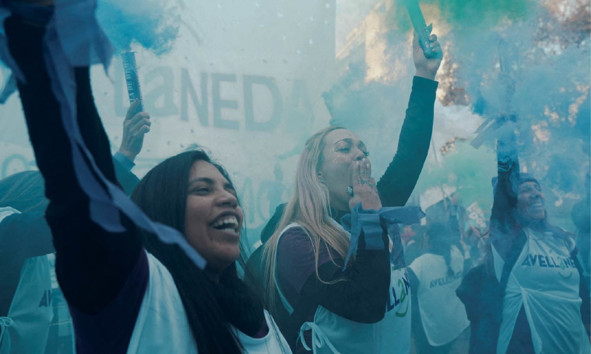 Two women at a protest for the ”Ni Una Menos movement raise their hands while holding cannon smoke powder poppers as blue powder surrounds them.