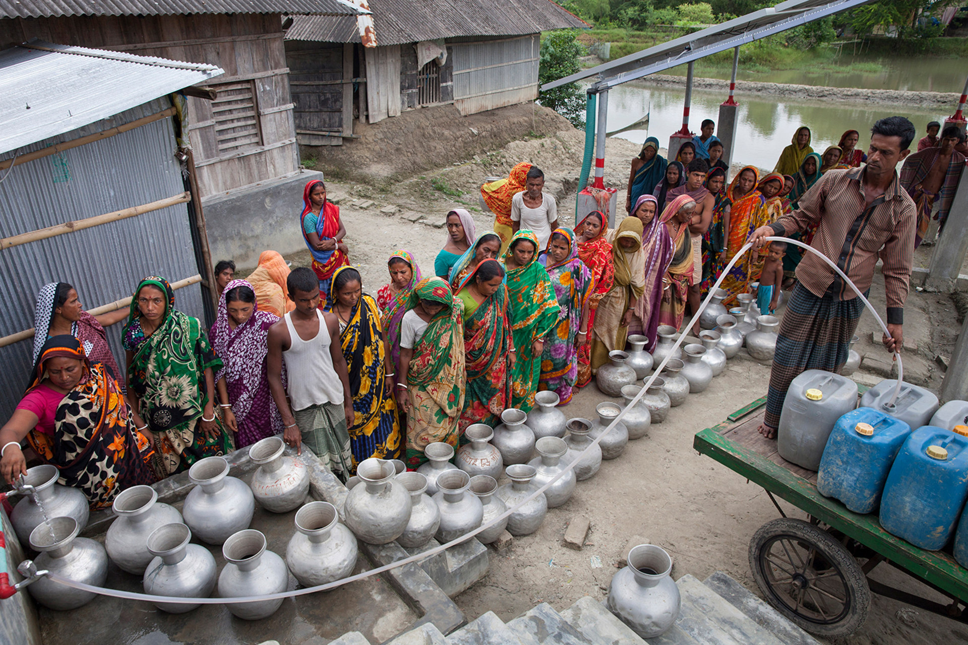 People stand in line to collect drinking water from a reverse-osmosis plant. There are multiple silver water containers waiting to be filled. A man on a cart fills water carriers using a pipe.
