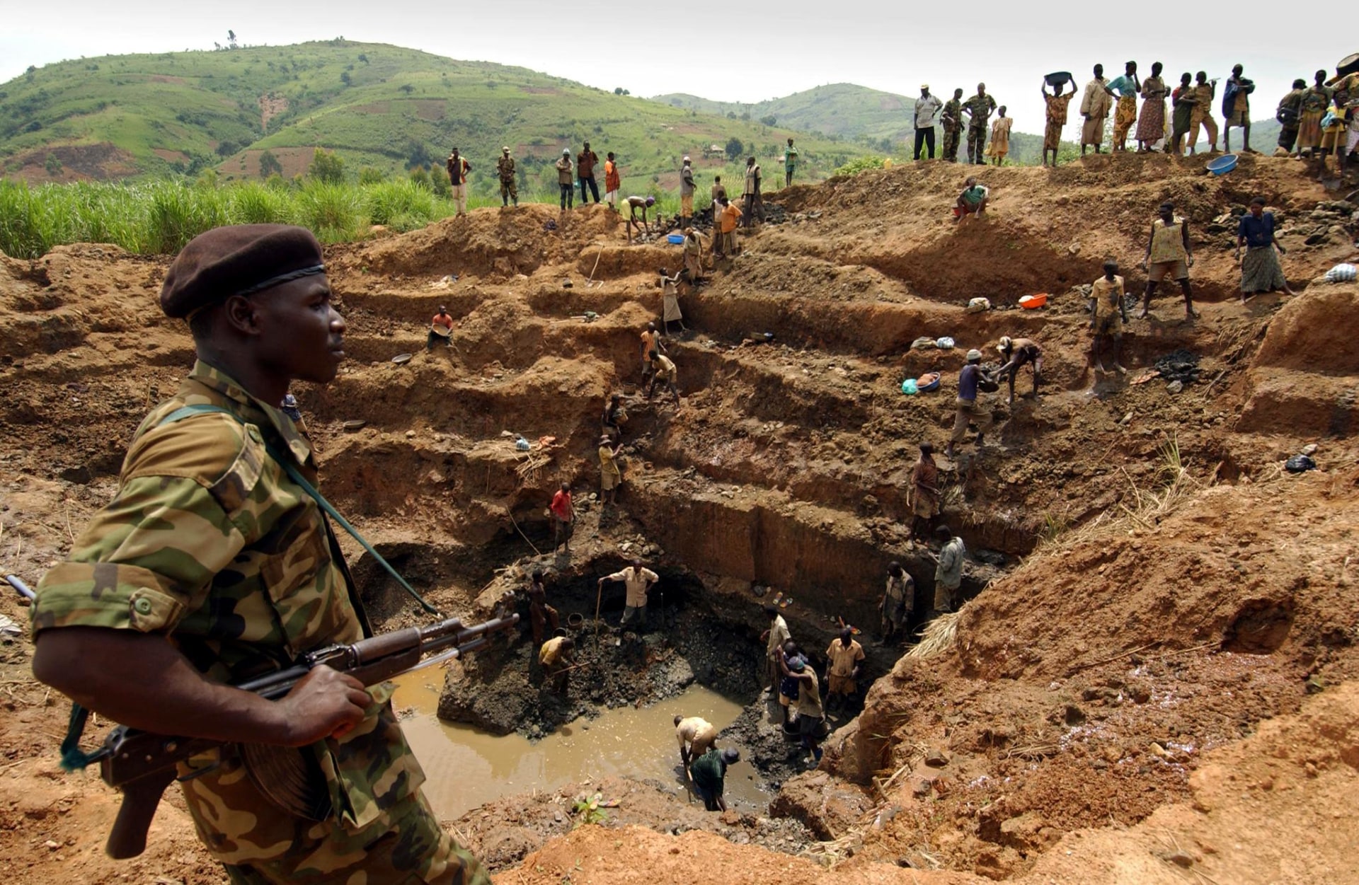Man in military-type uniform holds a machine gun while overlooking a mining operation with many laborers excavating mud in search of gold deposits.