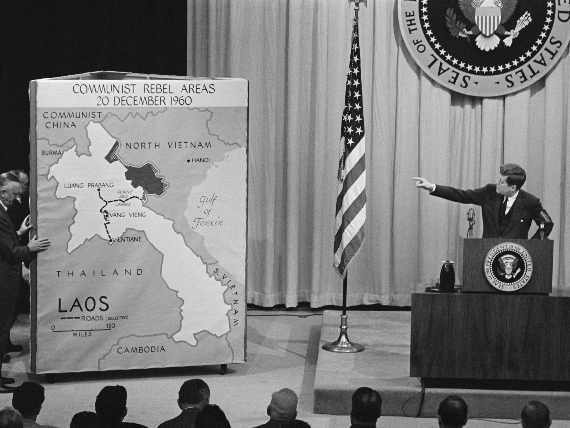 President John F. Kennedy, standing at a podium (on the righthand side), points leftward to a map of Laos while discussing the growth of Communism in southeast Asian countries at a news conference.
