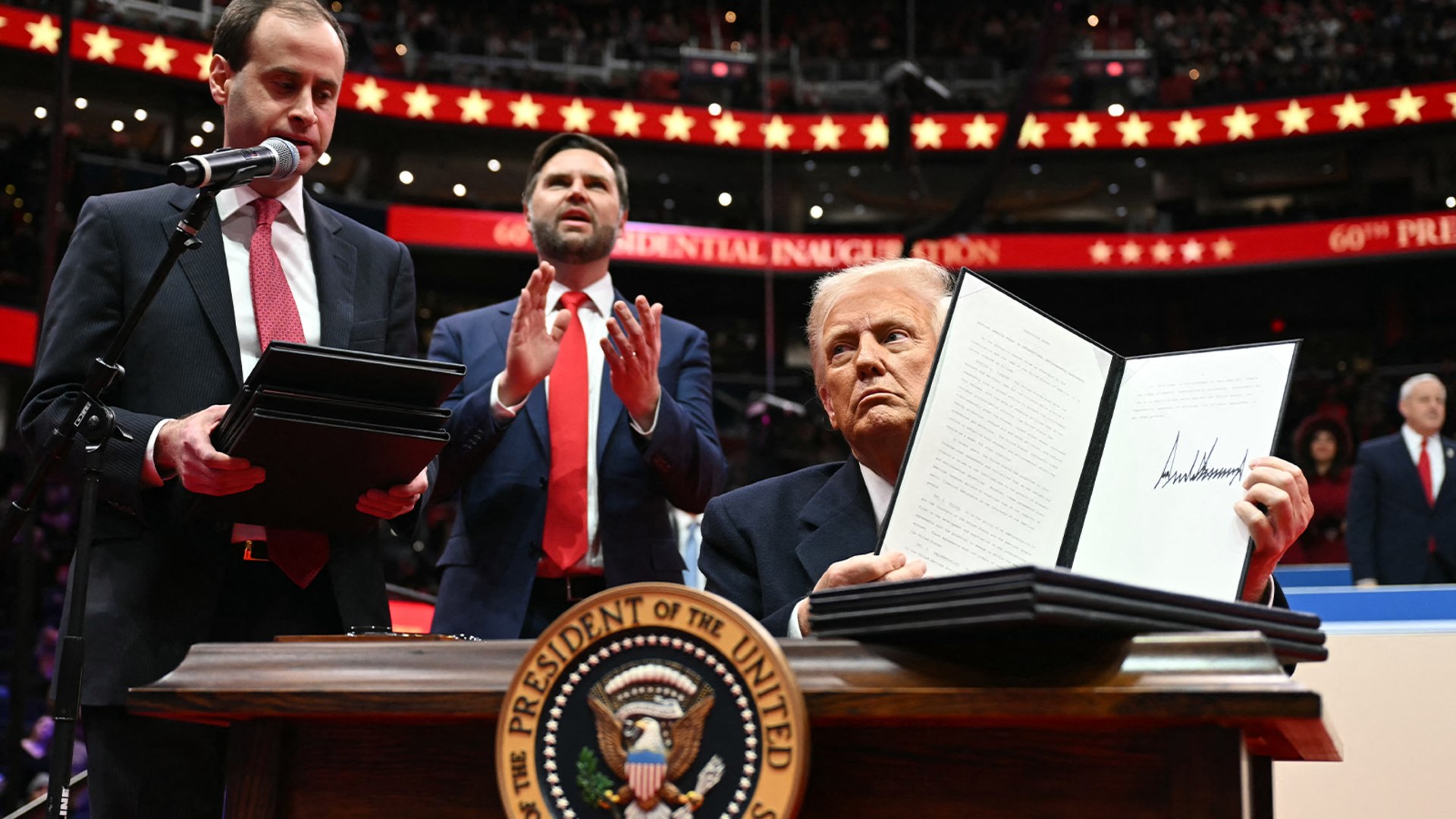 President Trump on a stage seated behind the seal of the United States holding up an executive order document with a large signature visible.