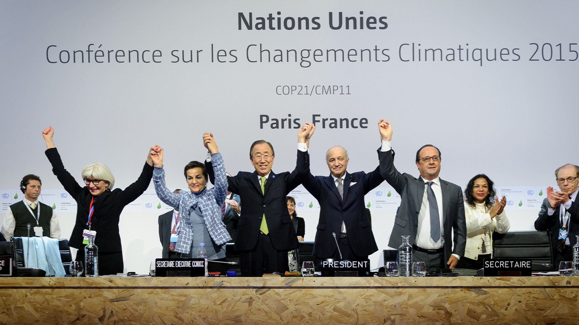 Officials in front of crowd raise arms together in celebration of the Paris Agreement.