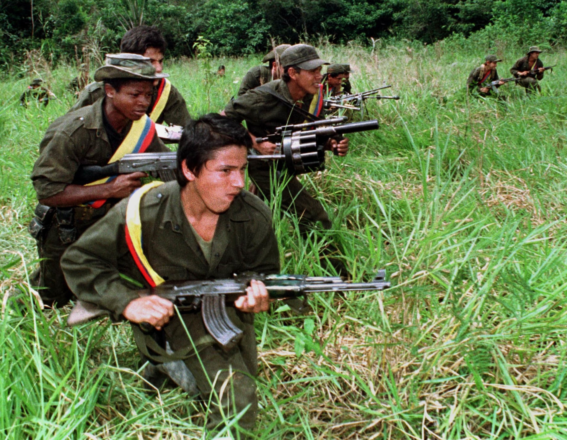 Guerrilla fighters of the Revolutionary Armed Forces of Colombia crouch in grass while patrolling the jungle with weapons.