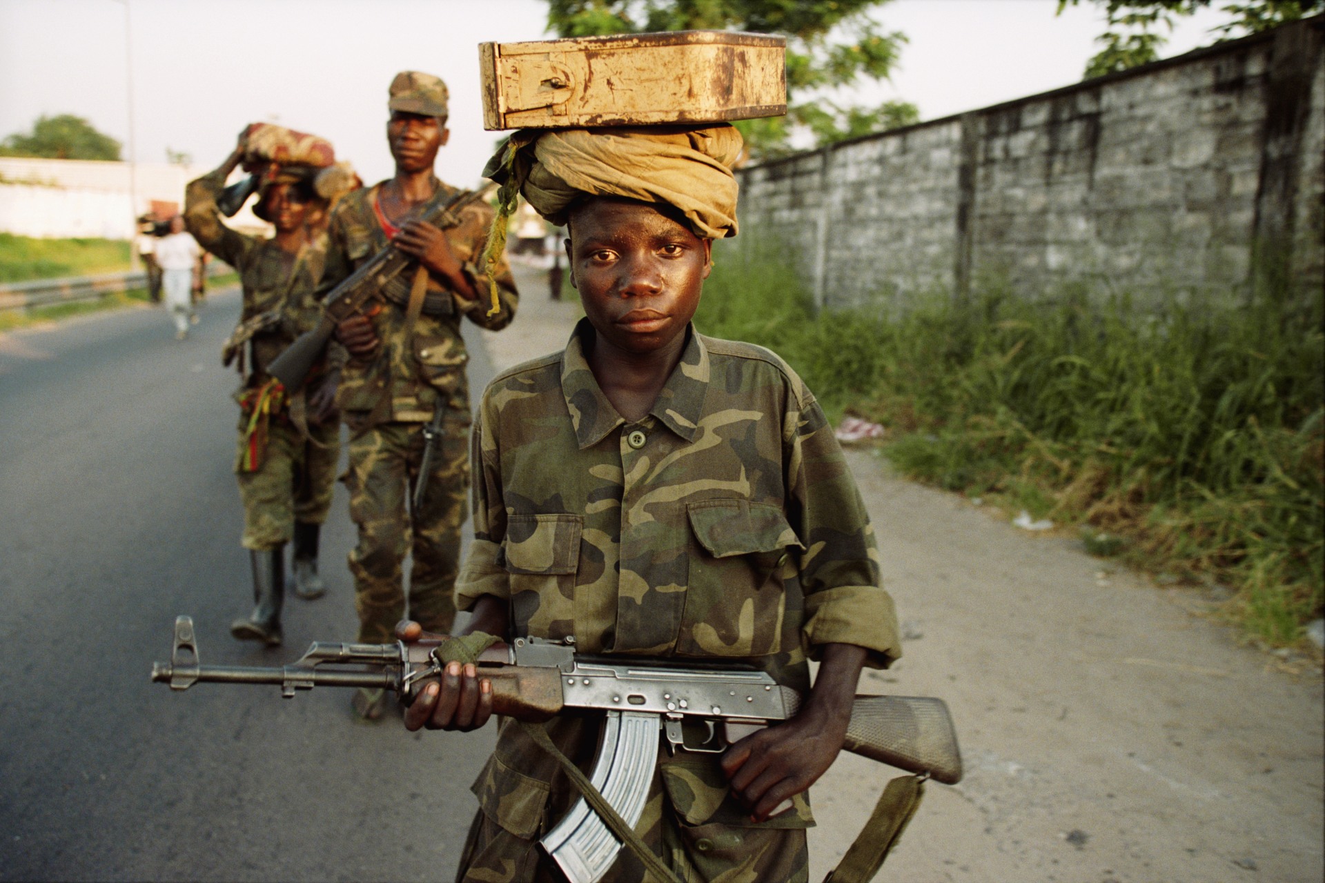 Child soldiers, wearing uniforms, hold guns and carry packages on their head.