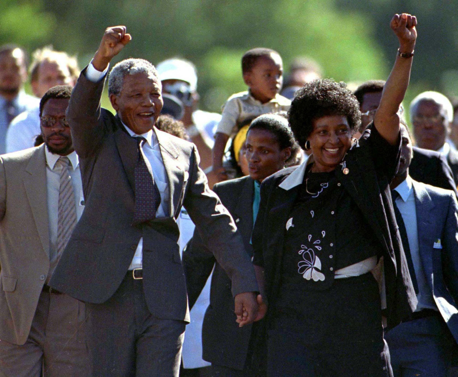 Nelson Mandela, accompanied by his wife Winnie, walks out of of prison. Both of them raise their fists in the air triumphantly as people walk behind them.