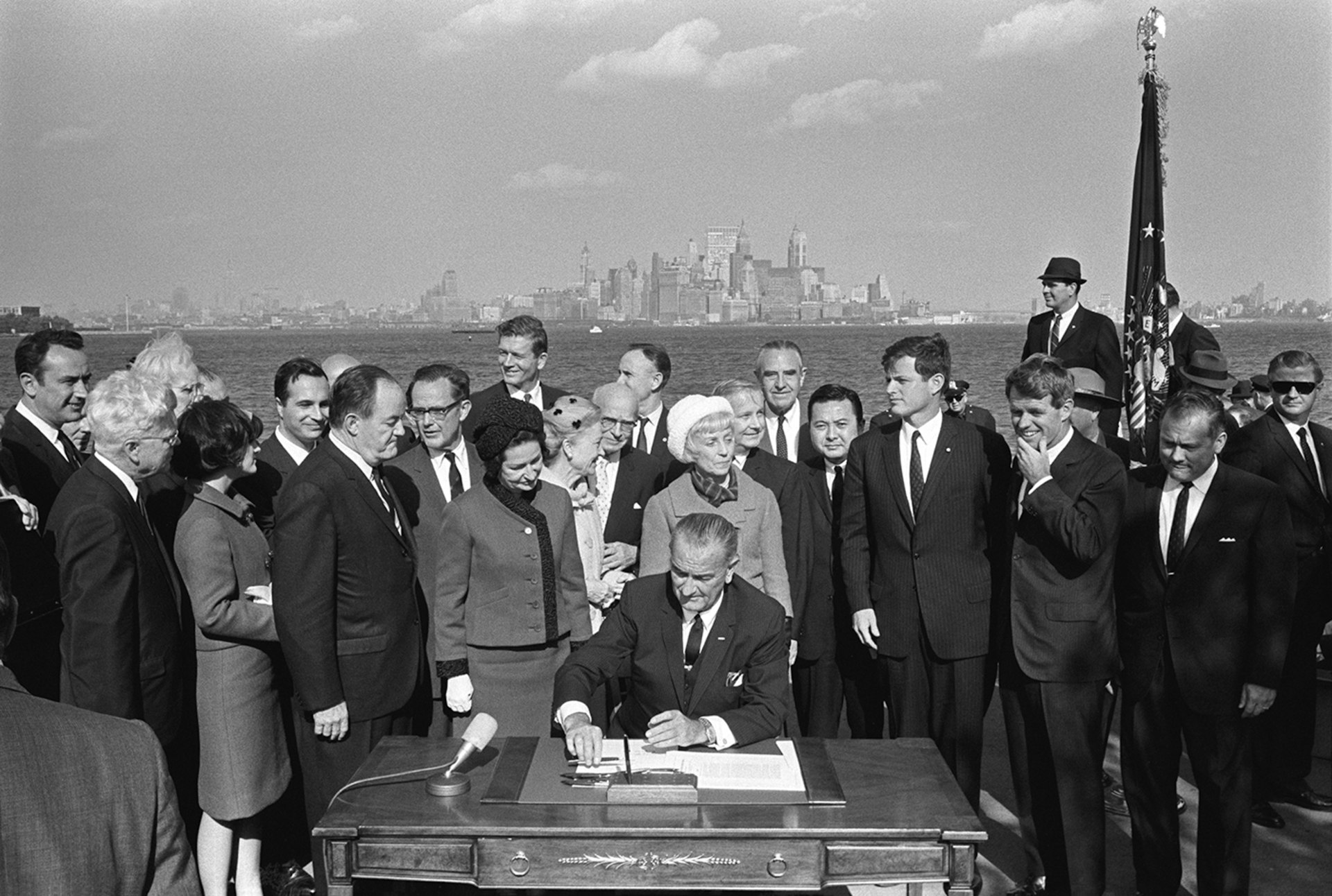 President Lyndon B. Johnson picks up a pen to sign papers while people gather around him and the New York City skyline shows in the background.