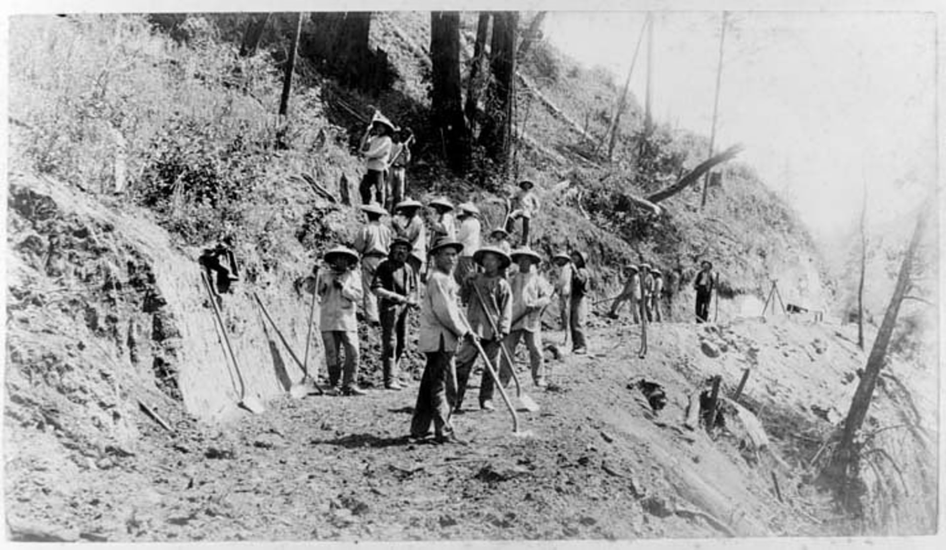 Chinese laborers holding shovels work on a railroad by a cliffside.