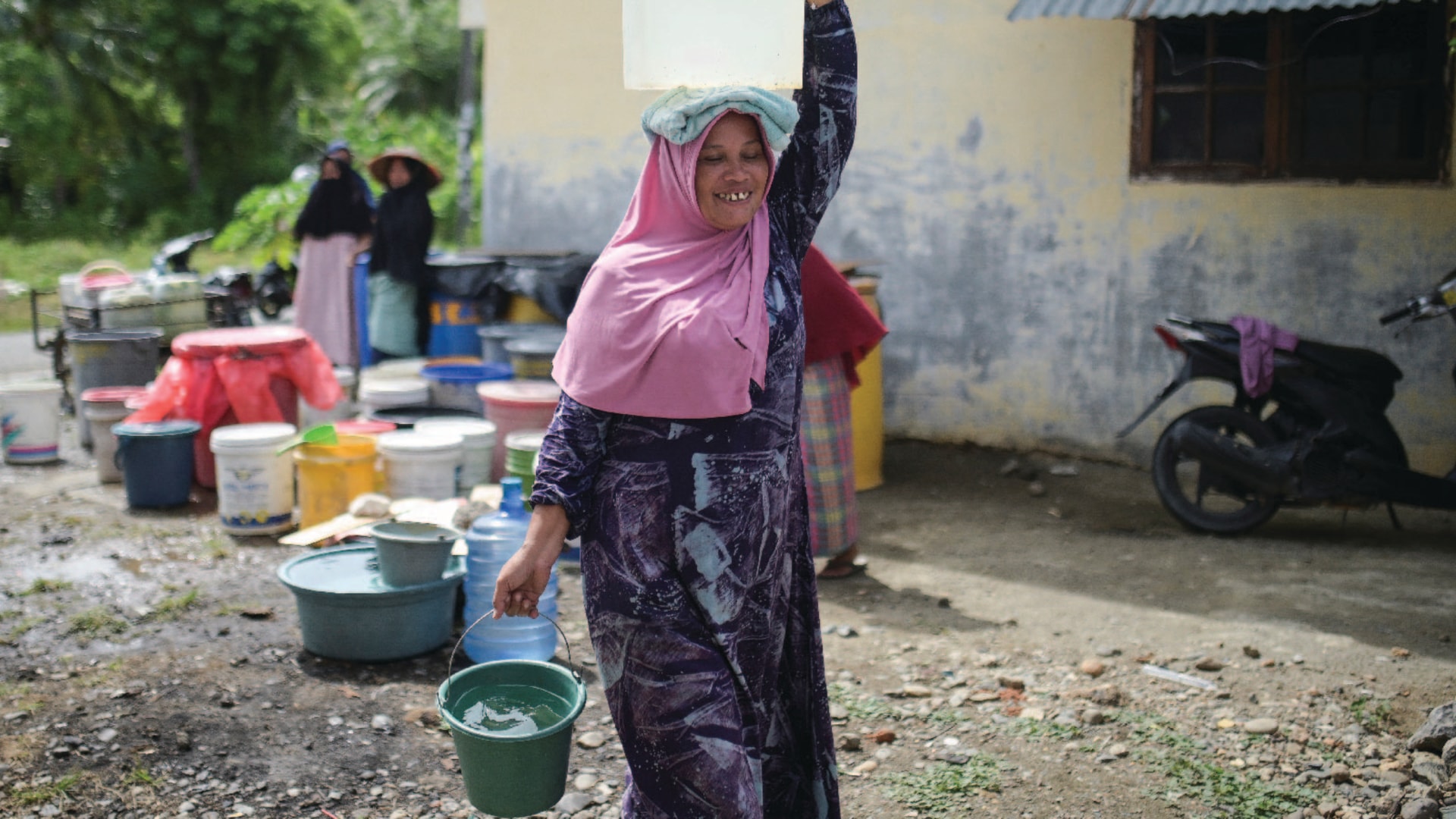 A woman carries a bucket of water in her hand and another jug of water on her head.
