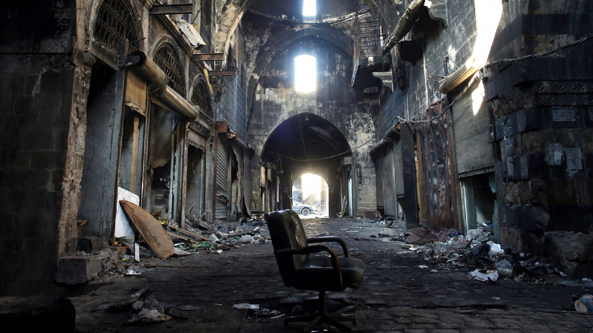 A destroyed Al-Madina Souq market in Aleppo. There is trash on the ground and a singular rolling chair can be seen in the center of the image.