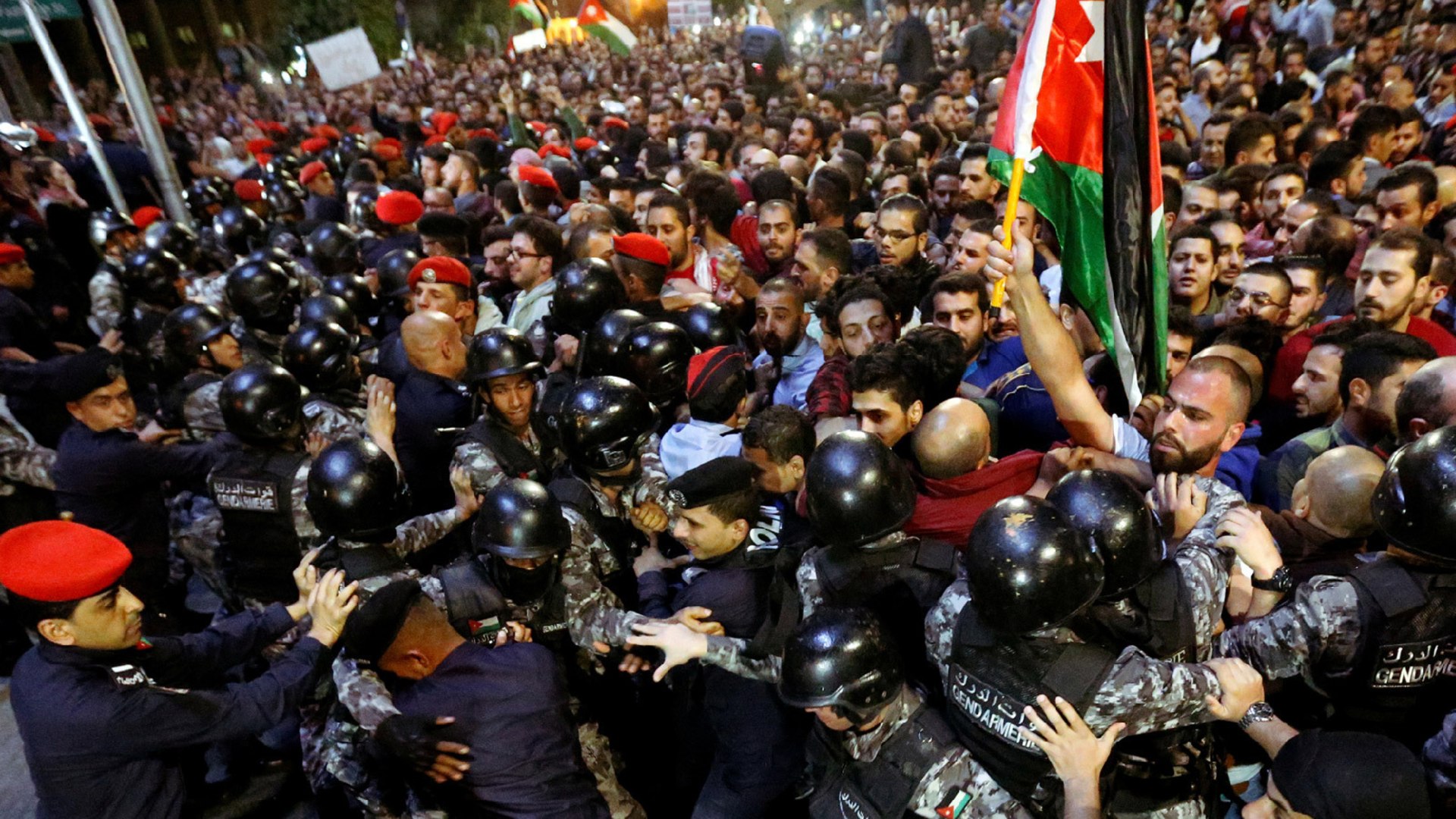 Policemen wearing black helmets and gear clash with a crowd protesting tax hikes in Amman, Jordan. Someone in the crowd holds up a Jordanian flag.