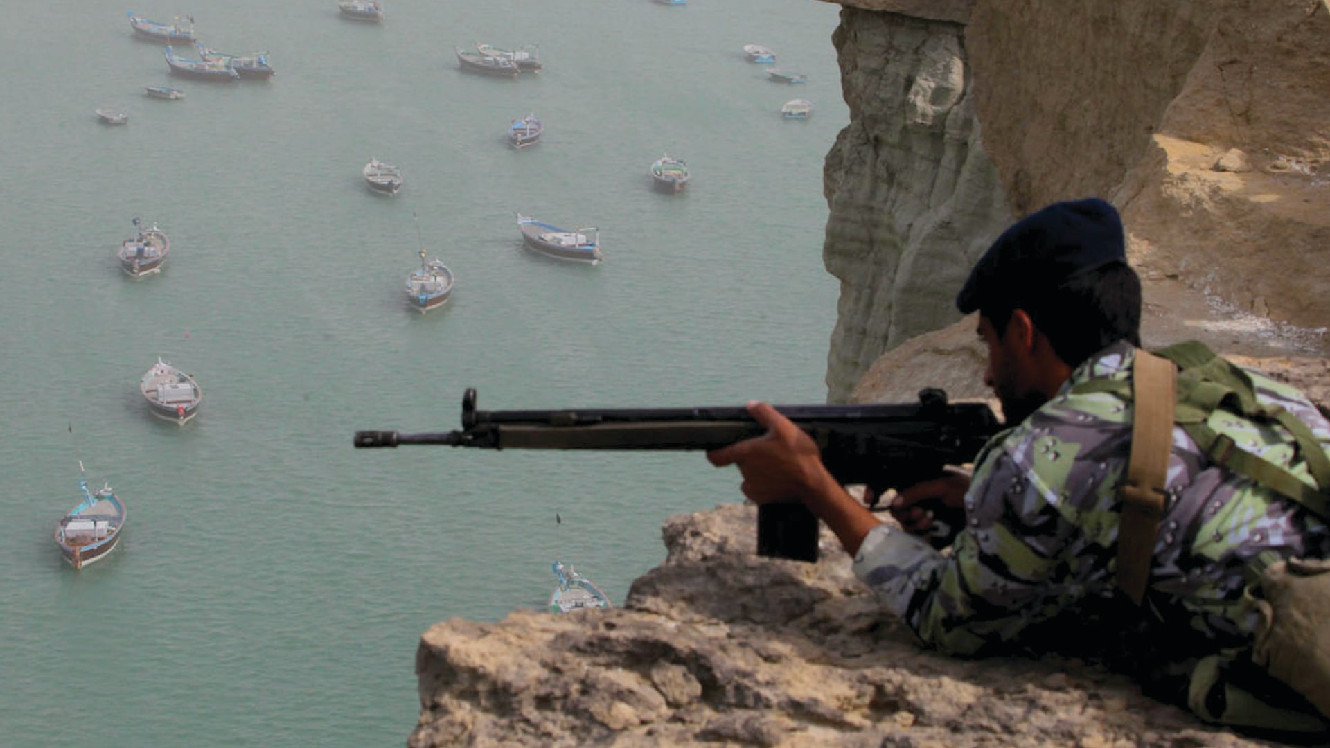 Iranian military personnel practice at rocky location near the Strait of Hormuz in southern Iran. Many boats can be seen floating in the water in the background.