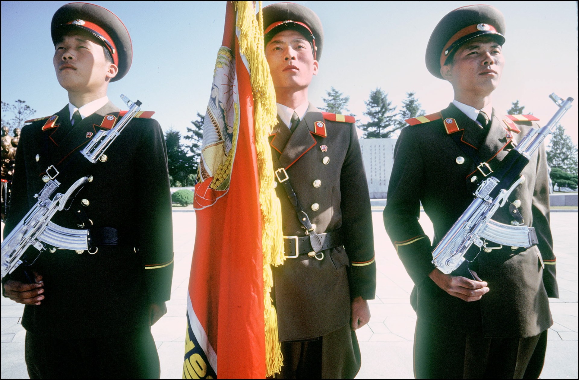 North Korean soldiers stand guard. The soldiers on the left and right are holding guns, while the soldier in the center is holding a flag.